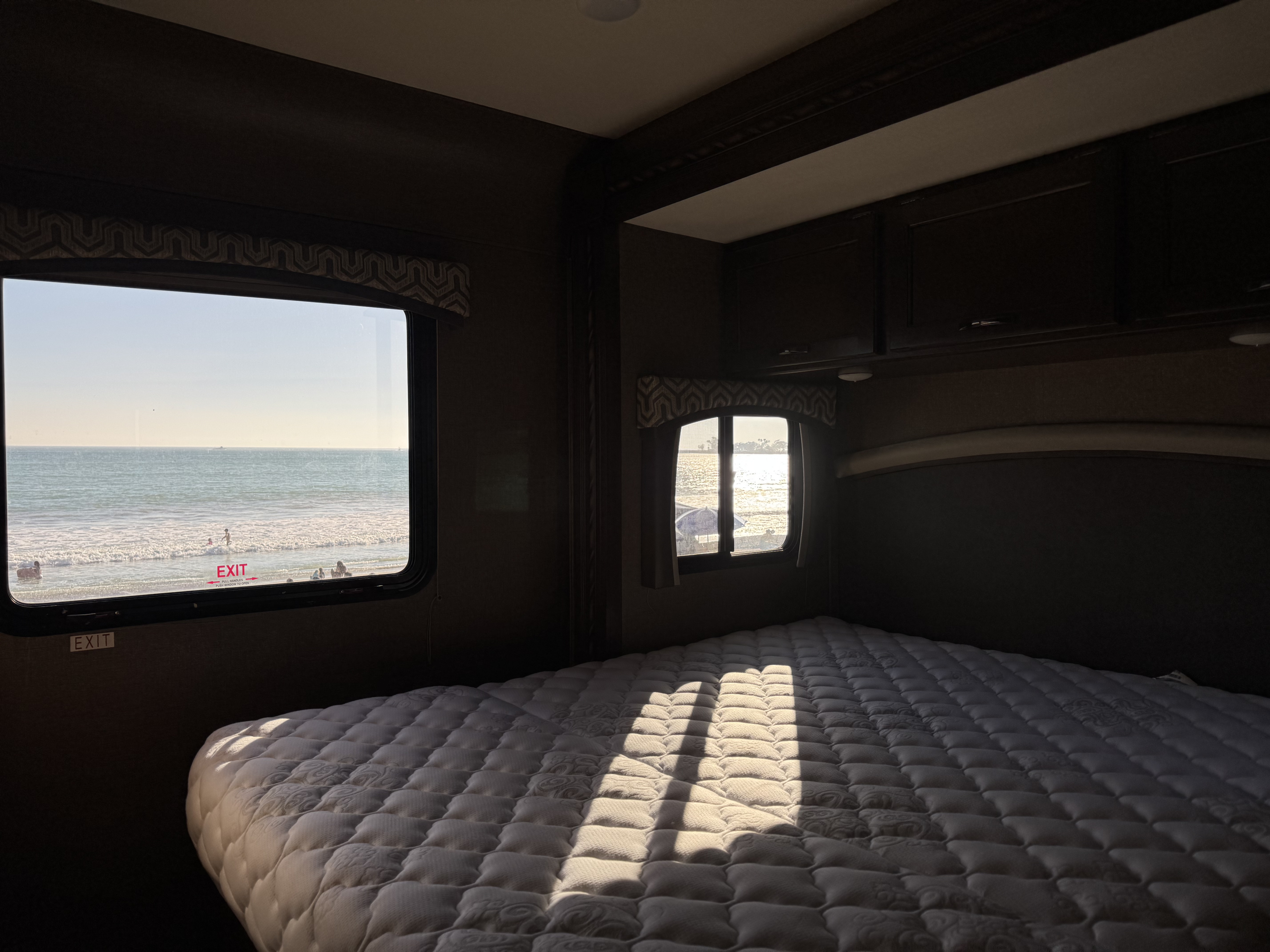 Bright morning in an RV rental bedroom overlooking the ocean. Sunbeams highlight the quilted bed, with beachgoers outside.. Thor Chateau 2019