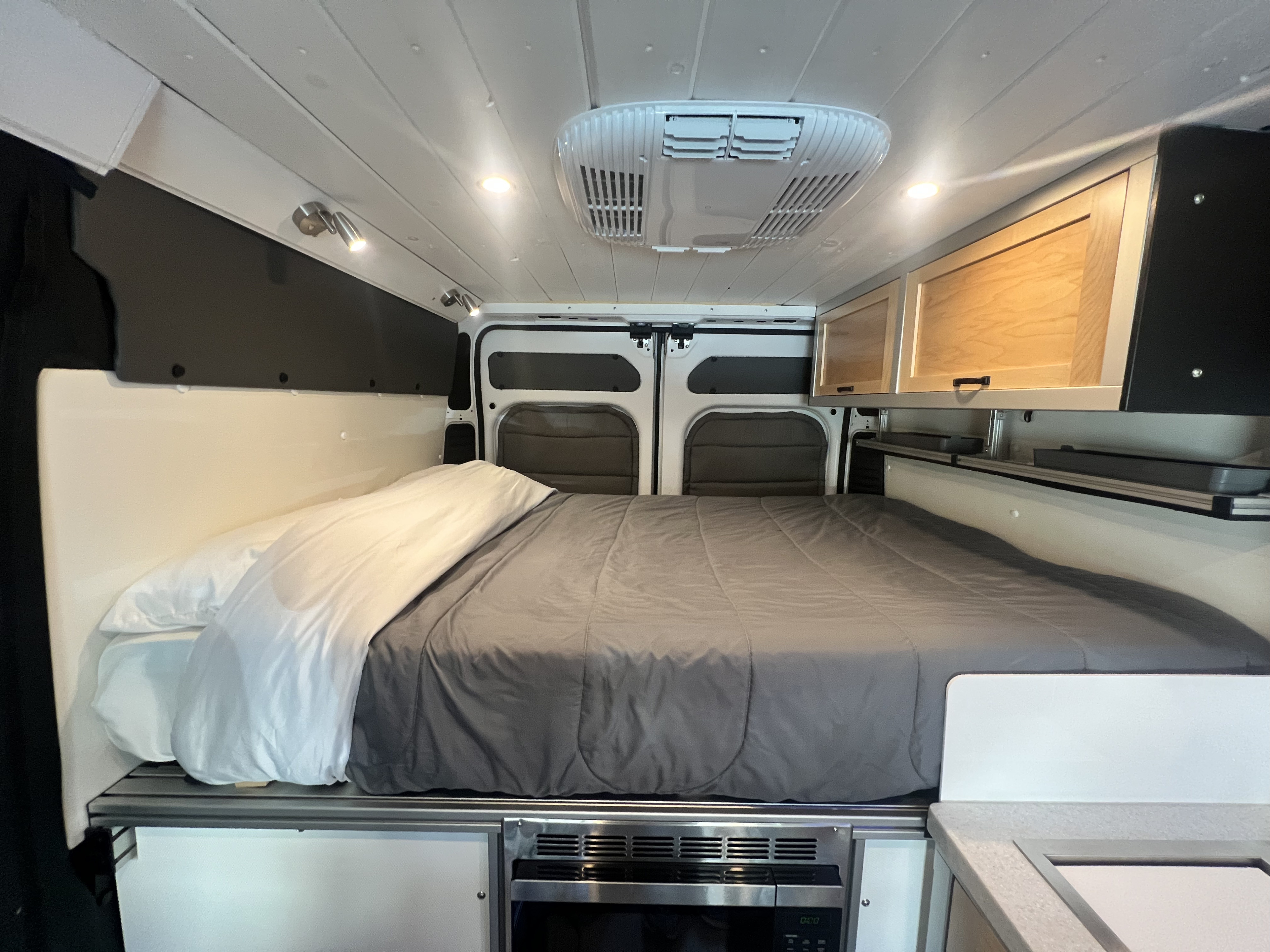 This cozy RV rental interior features a queen-sized bed, overhead storage cabinets, and a convenient under-bed microwave.. Ram Promaster 1500 2020