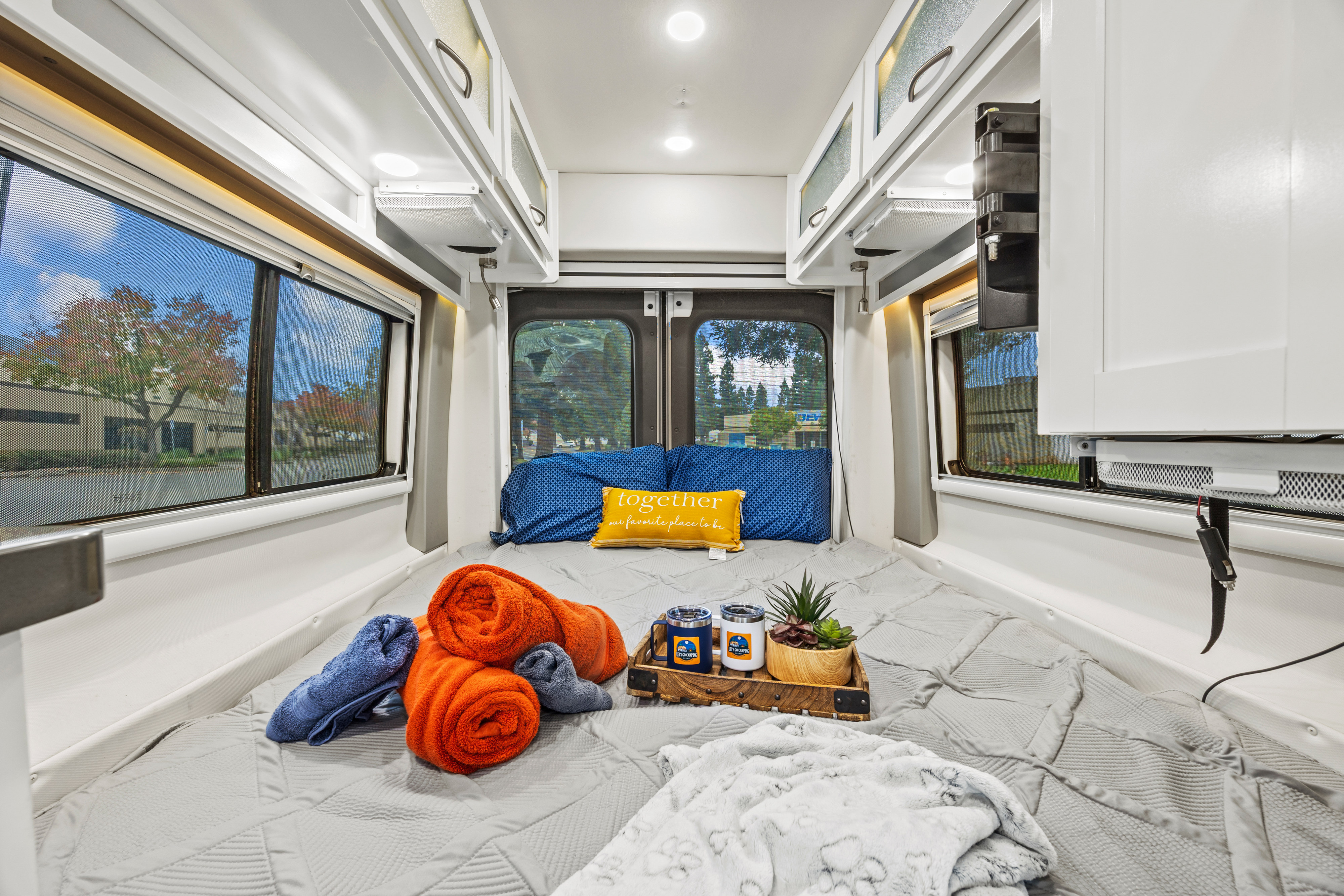**rv rental**: A cozy bed with pillows, rolled towels, and a plant on a tray. Overhead cabinets, bright lighting, and screened windows offer comfort.. Coachmen Beyond 2021
