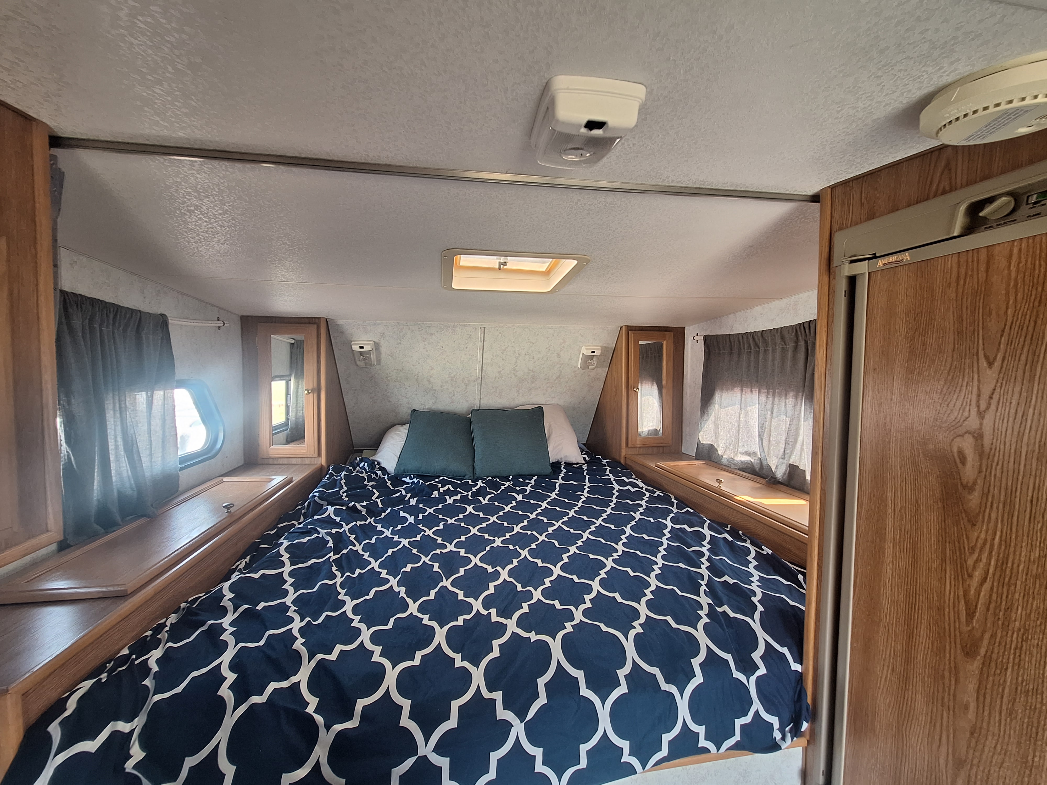 rv rental: Inside a compact bedroom, a navy patterned bed, side storage, windows with curtains, and a fridge.. Glacier Campers Truck Camper 2016