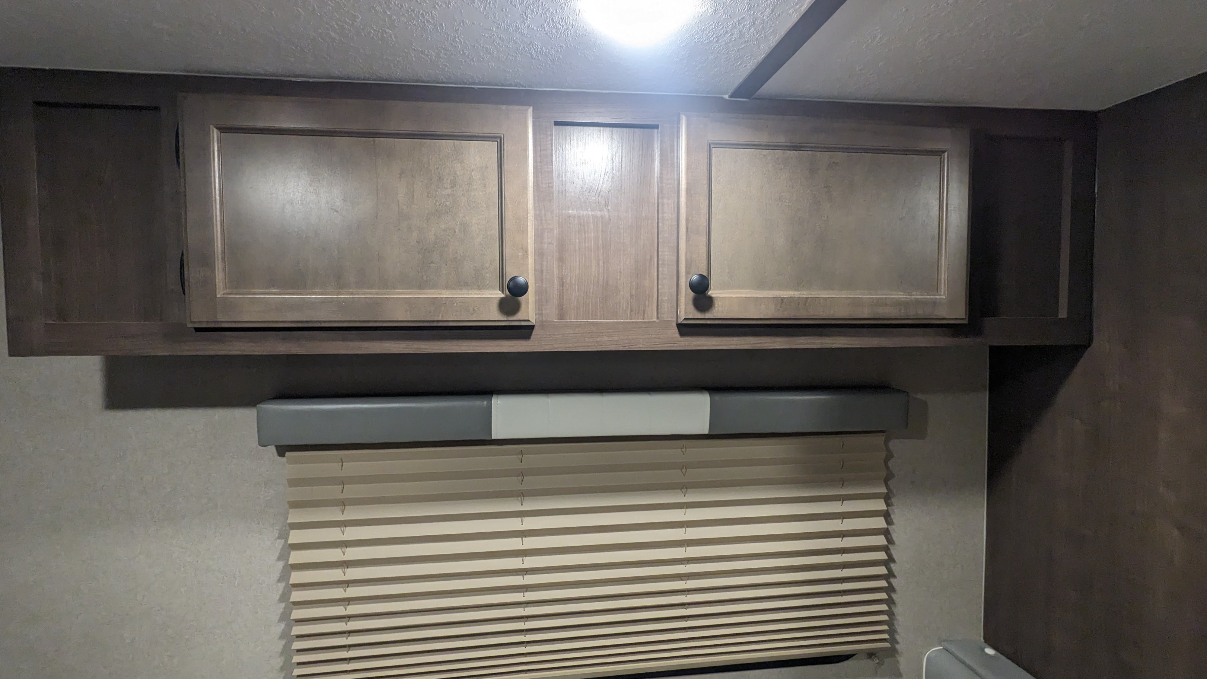 Inside an RV rental: overhead wood cabinets with black knobs and a pleated window blind for storage and privacy.. Sunset Park RV Sun Lite 2022
