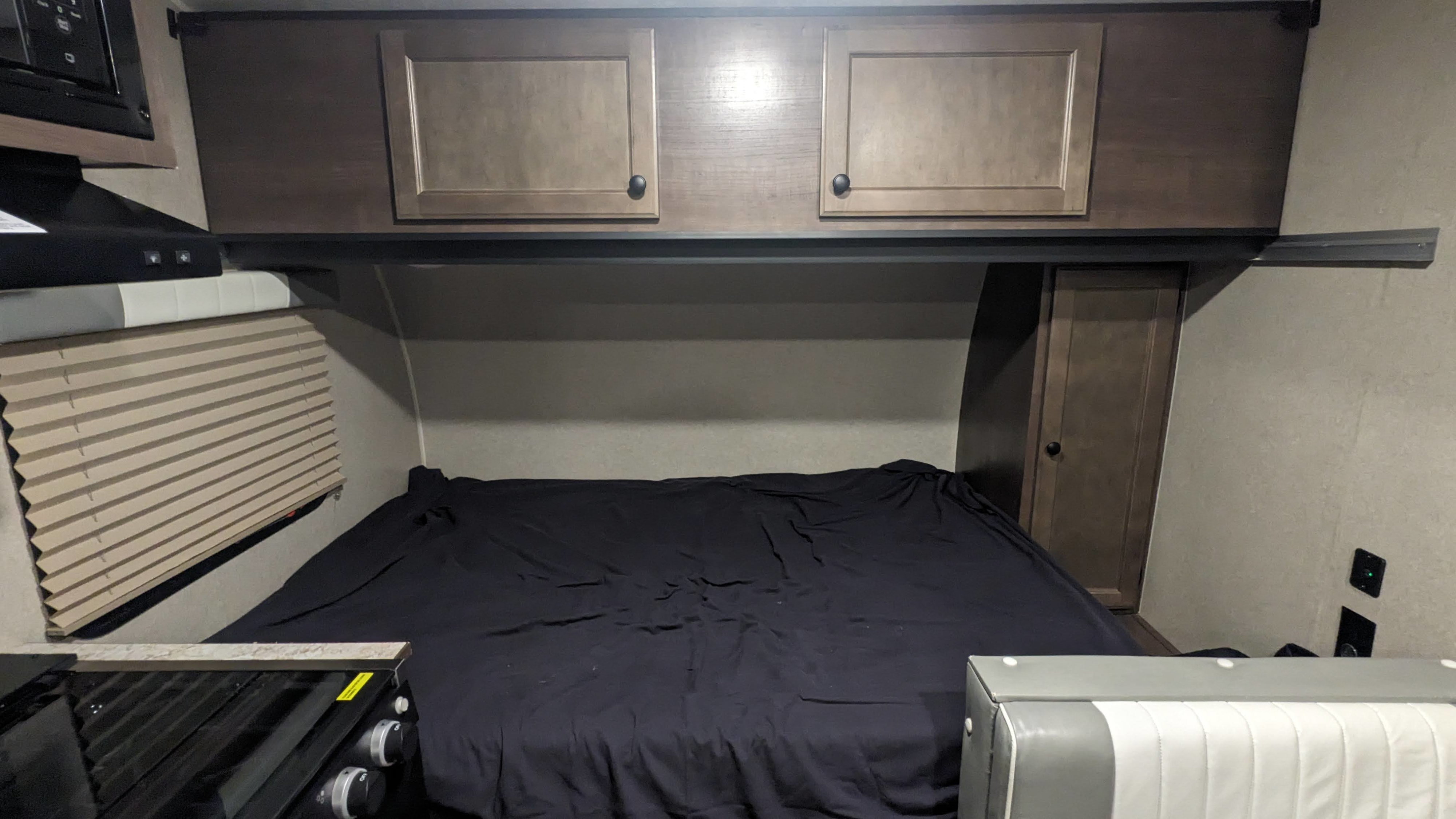 Cozy rv rental interior showing a comfortable bed with dark covers, overhead cabinets, and a compact kitchen area.. Sunset Park RV Sun Lite 2022