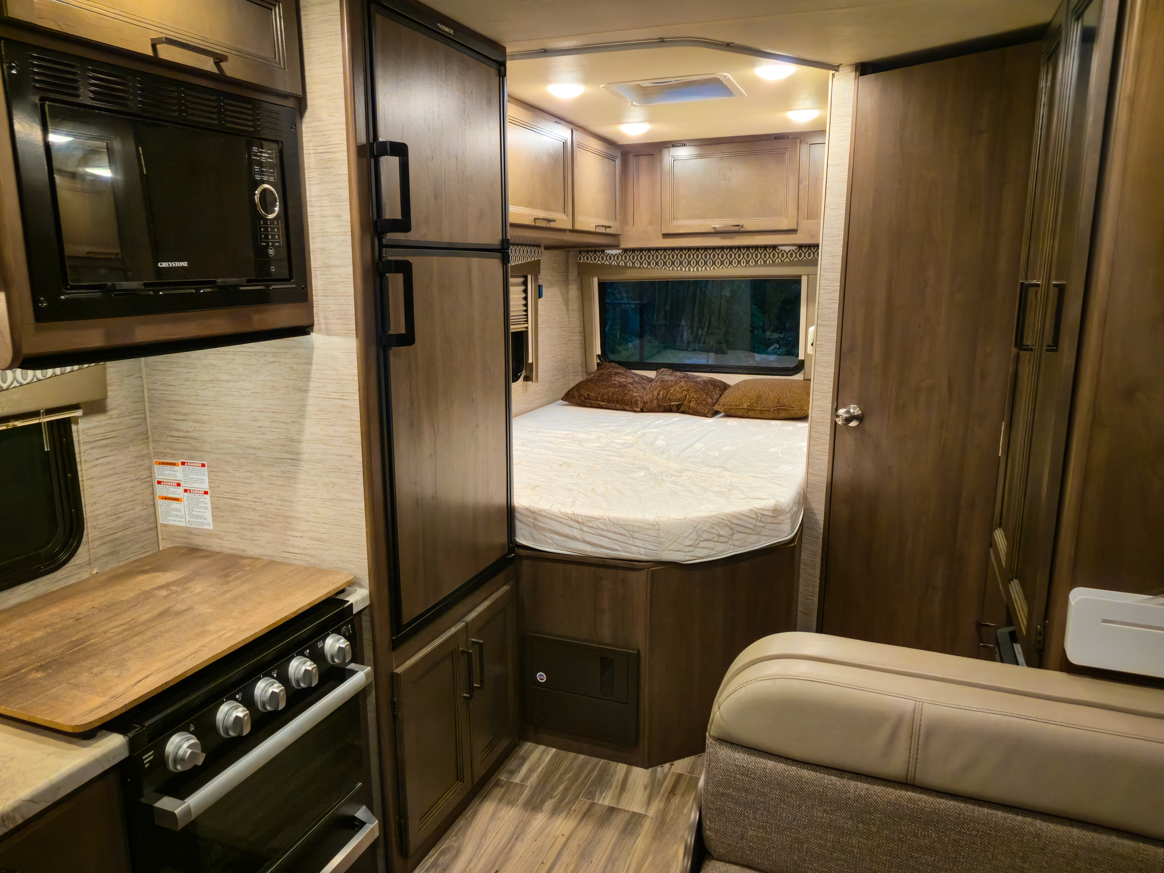 Compact rv rental interior: microwave, stove, fridge, and a bed with window visible. Warm wood finishes create a cozy space.. Thor Freedom Elite 2021