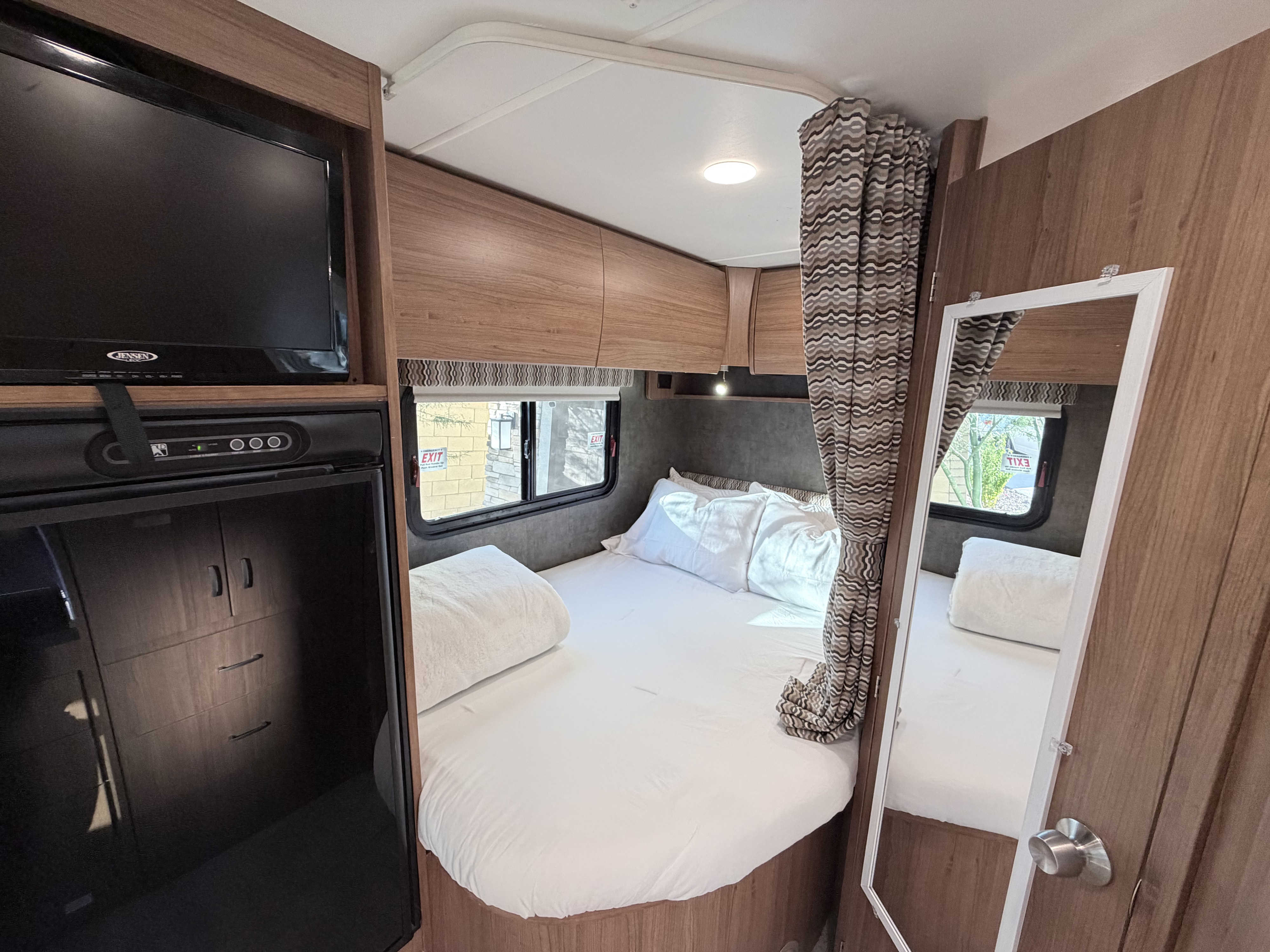 Clean RV rental bedroom featuring a white bed, TV, storage cabinets, and mirror. Ideal for compact living.. Winnebago Trend 2015