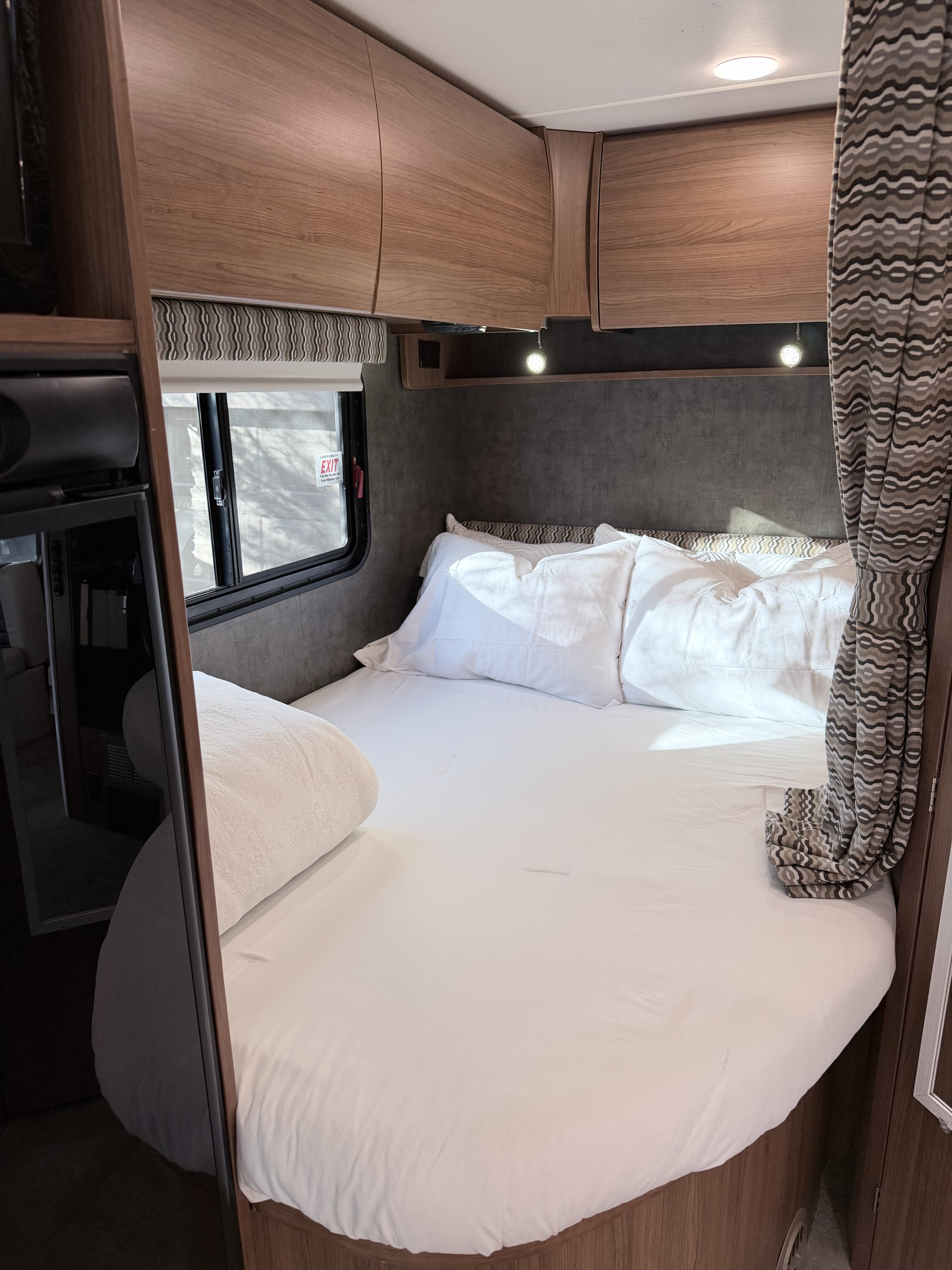 Inside a cozy RV rental: a neatly made bed with white linens, wooden overhead cabinets, and a window with a blind.. Winnebago Trend 2015