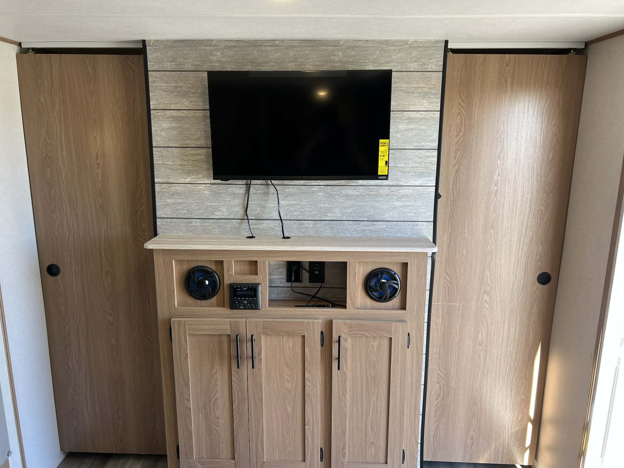 This modern rv rental features a central entertainment unit with a TV, built-in speakers, and storage cabinets, flanked by two large wooden closet doors.. AVENGER 24BHSLE 2023