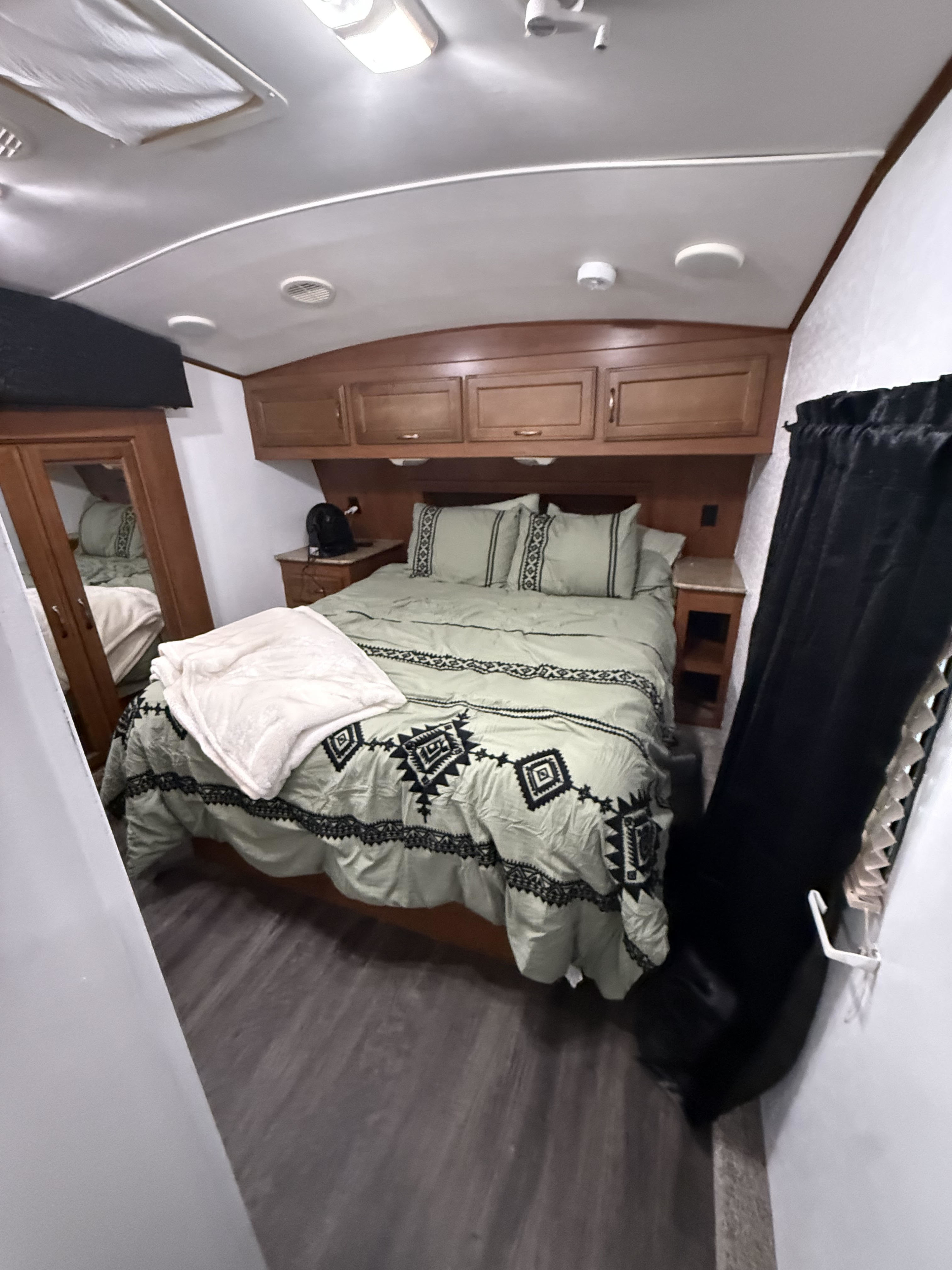 RV rental bedroom: light green bed with geometric patterns, wooden cabinets, nightstands, mirror, black curtains, and ceiling vents.. Heartland RVs Sundance 2013