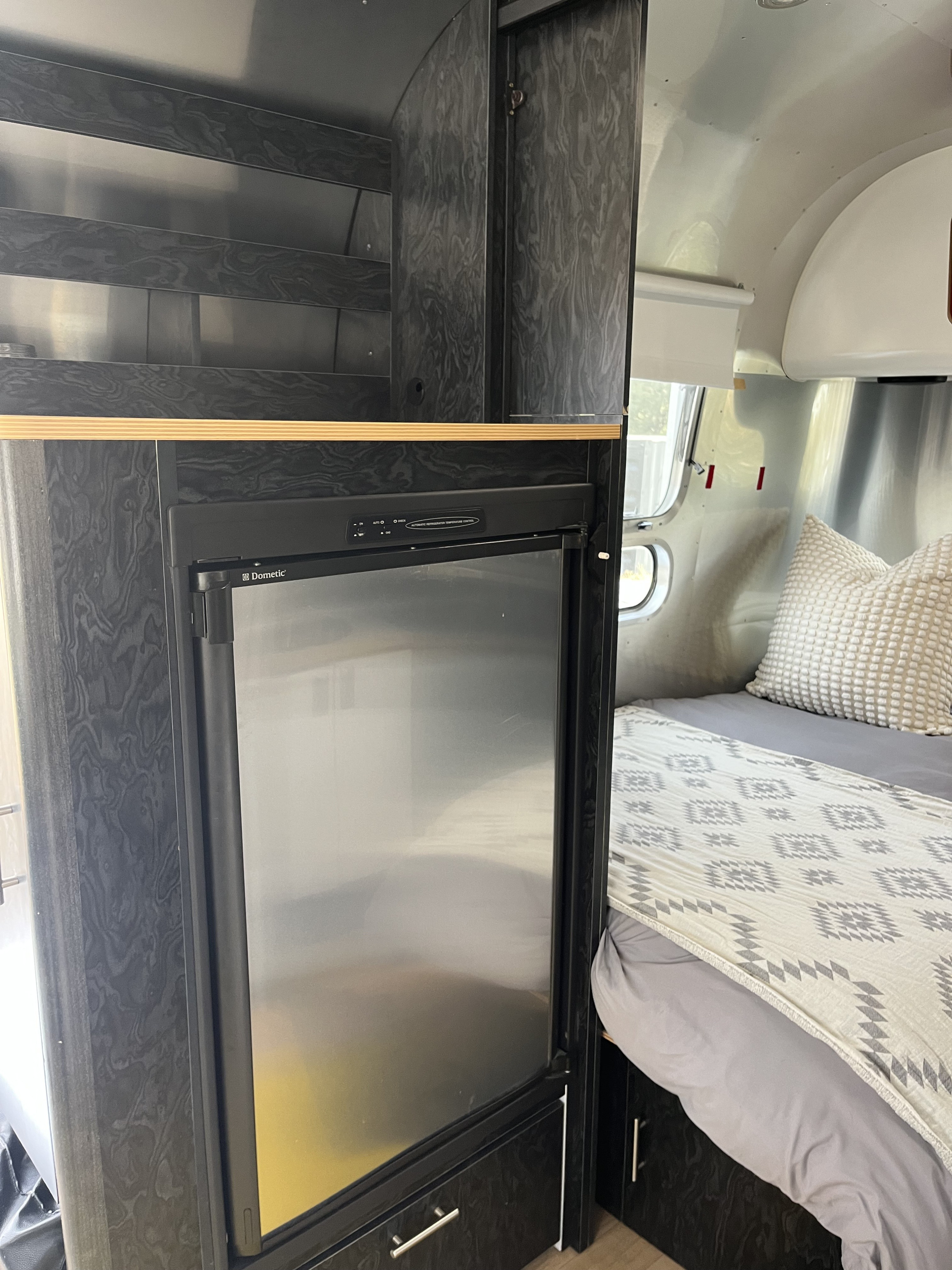Compact RV rental interior featuring a Dometic fridge, dark wood-look cabinetry, and a cozy bed area with a patterned blanket.. Airstream Bambi International 2006