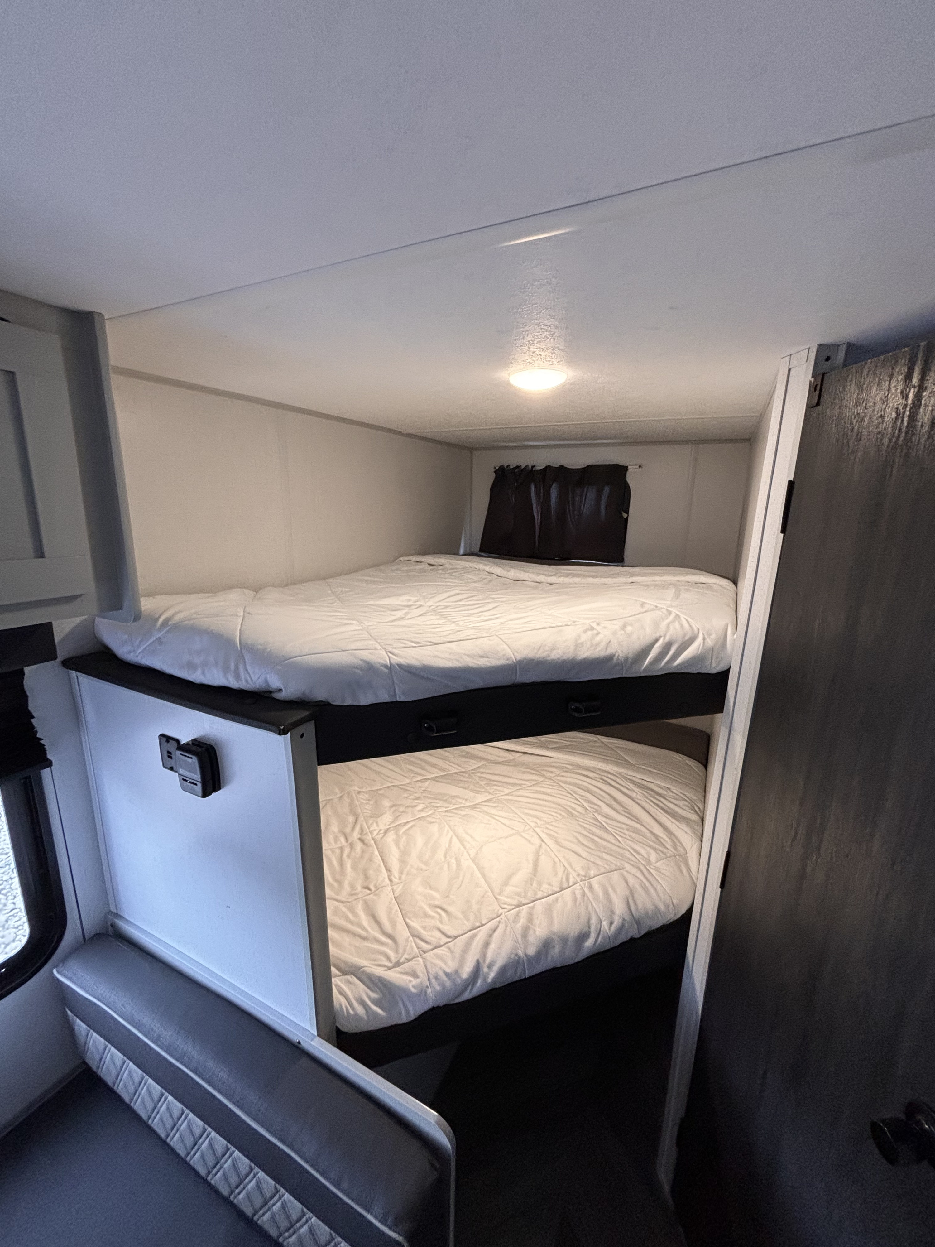 RV rental interior featuring two comfortable bunk beds with white comforters, a window with a black curtain, and a small bench seat.. Forest River Wildwood FSX Plantium 2022