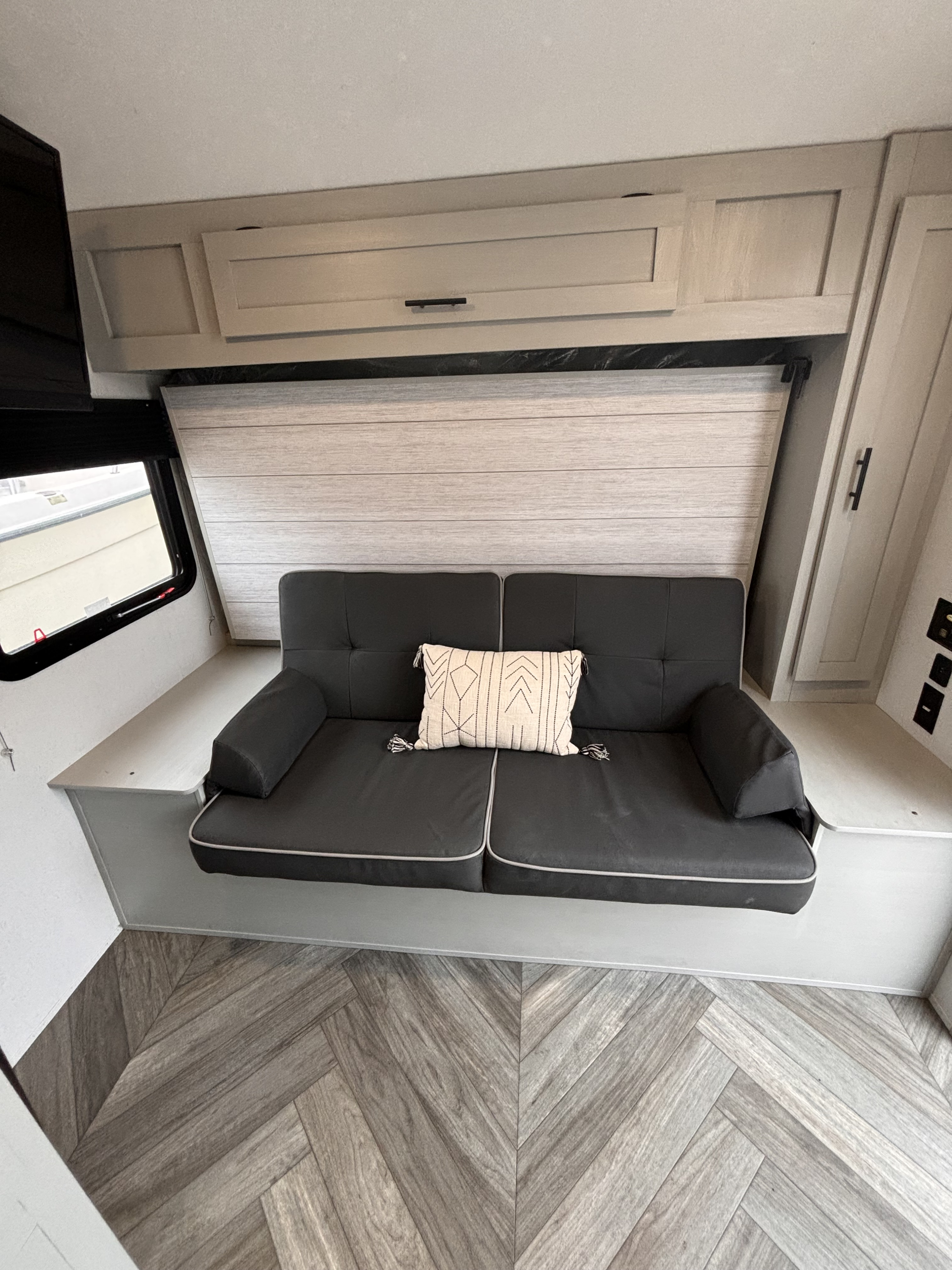 Step into this modern RV rental featuring a dark gray convertible sofa, overhead cabinets, and herringbone floor.. Forest River Wildwood FSX Plantium 2022