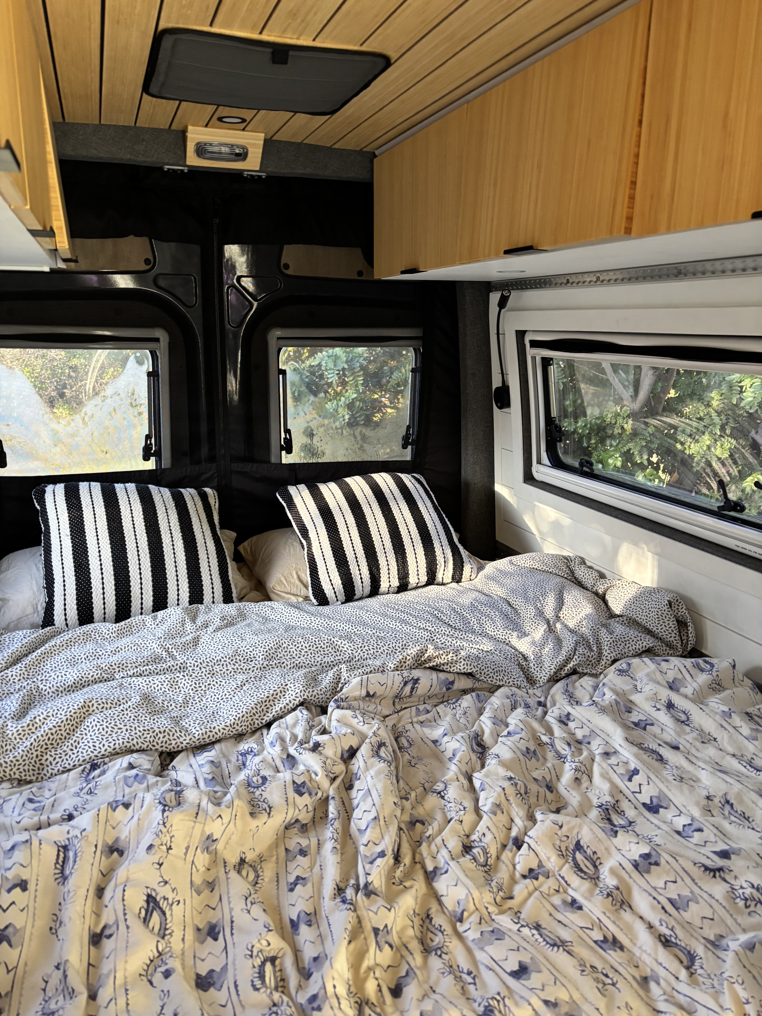 Inside an rv rental: A cozy bed with striped pillows, patterned bedding, and scenic nature views from the windows.. Mercedes Sprinter 2022