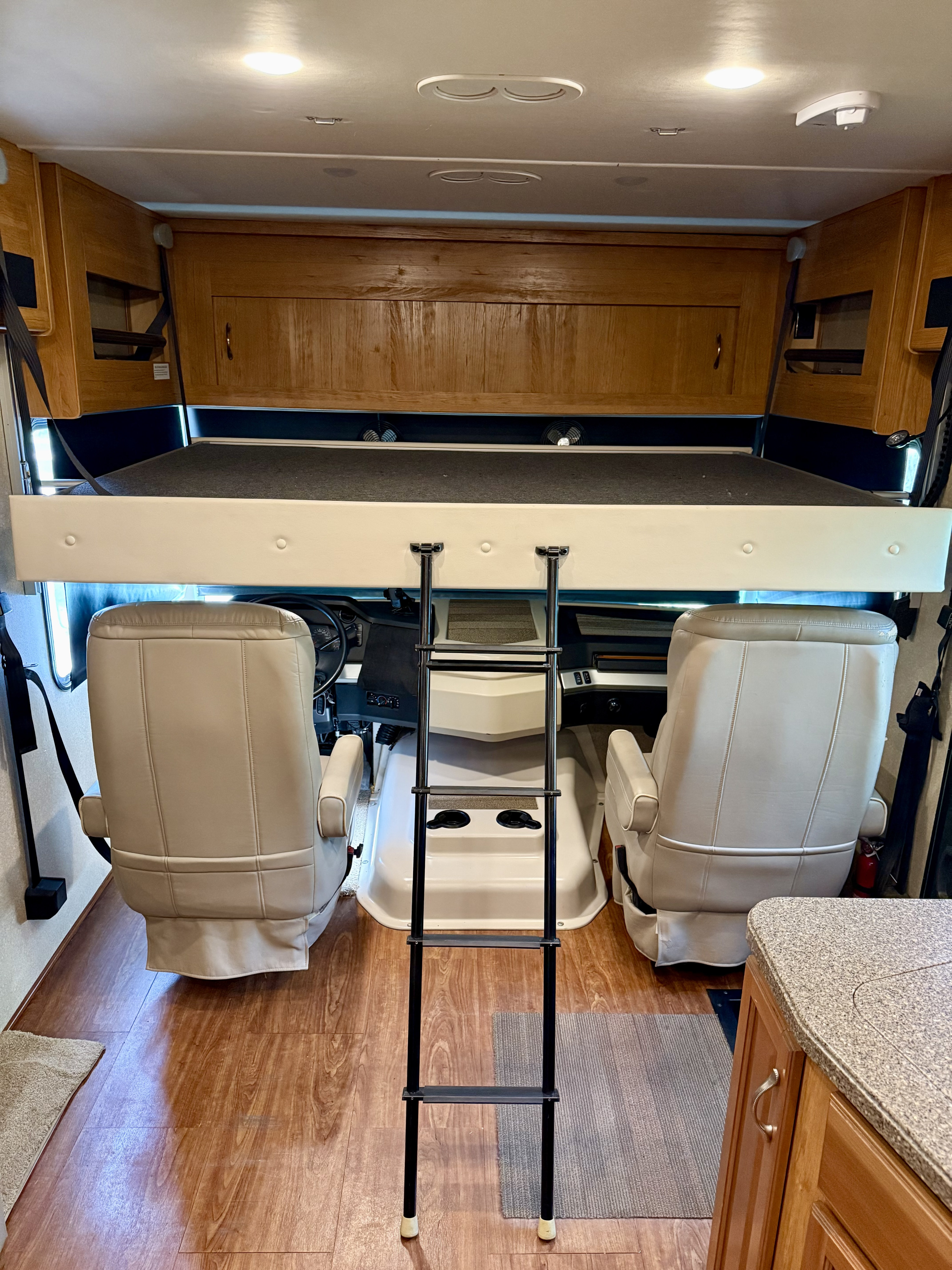 Spacious RV rental interior: cockpit with two plush seats, overhead bunk, and a ladder for access. Features wood trim & flooring.. Fleetwood Bounder 33C 2016