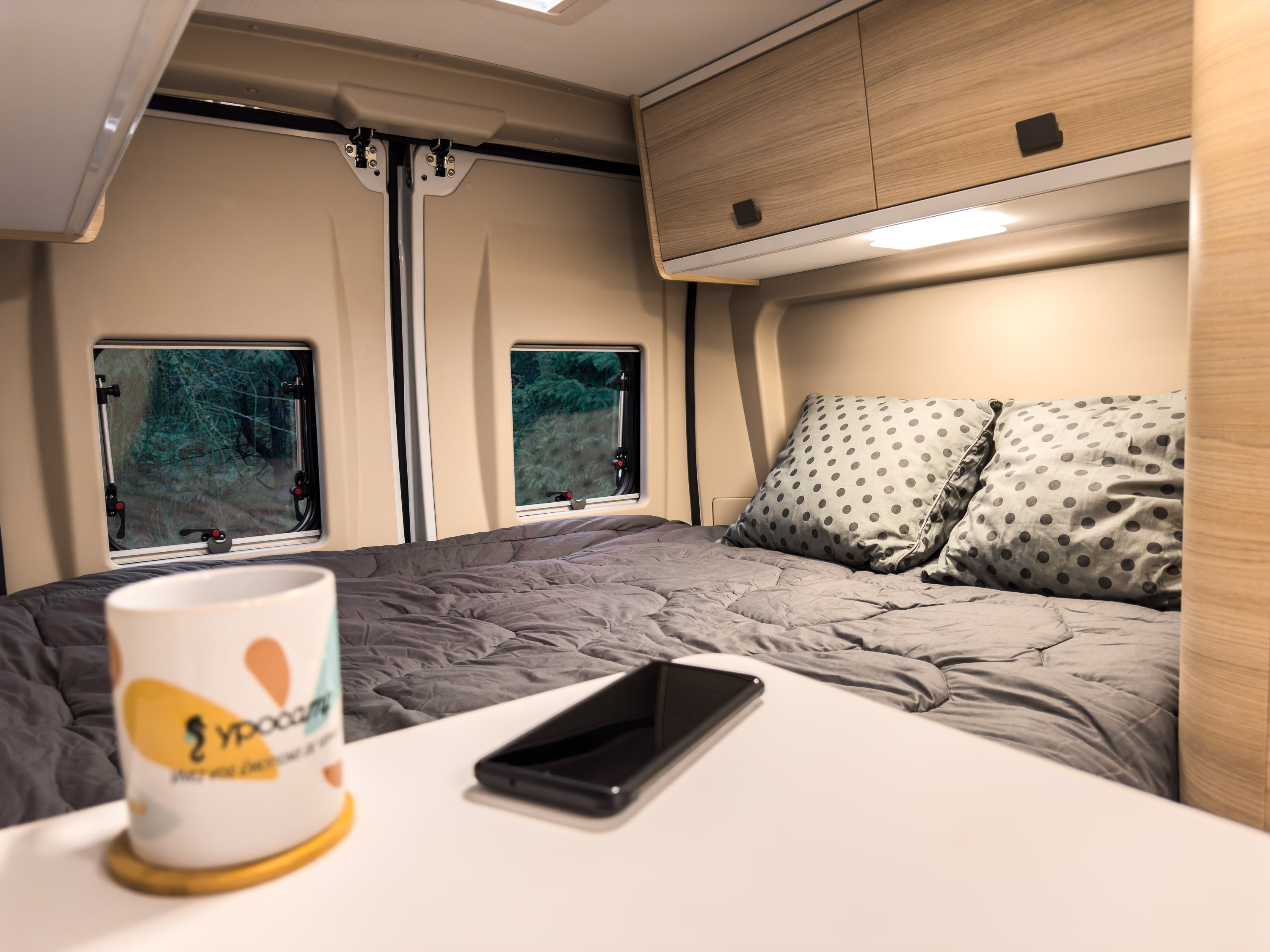 Cozy camper van RV rental interior: a comfortable bed with pillows, forest views from windows, a mug, and phone on the table.. Adria Twin sport 600 SX 2023
