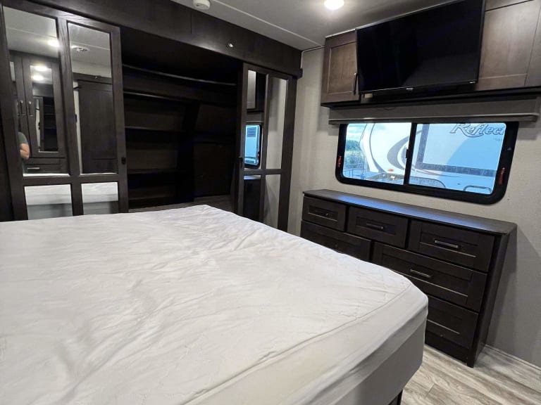 RV rental bedroom with a white bed, mirrored wardrobe to the left, and a dresser & TV opposite, above a window.. Grand Design Reflection 2020