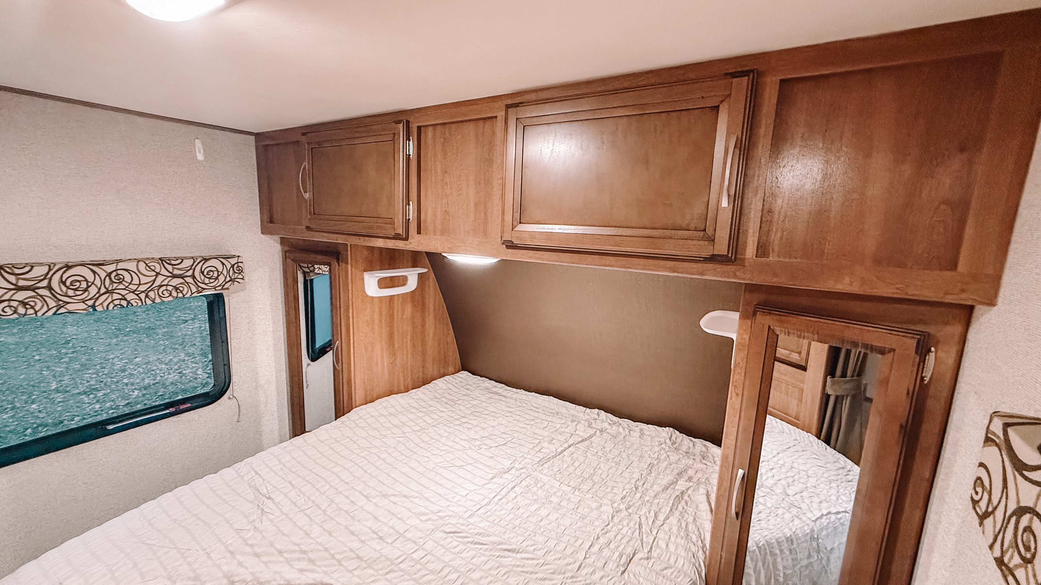 RV rental bedroom interior: light bedding, wood cabinets, small window, and a wall-mounted mirror. Cozy and compact space.. Coachmen Apex 2016