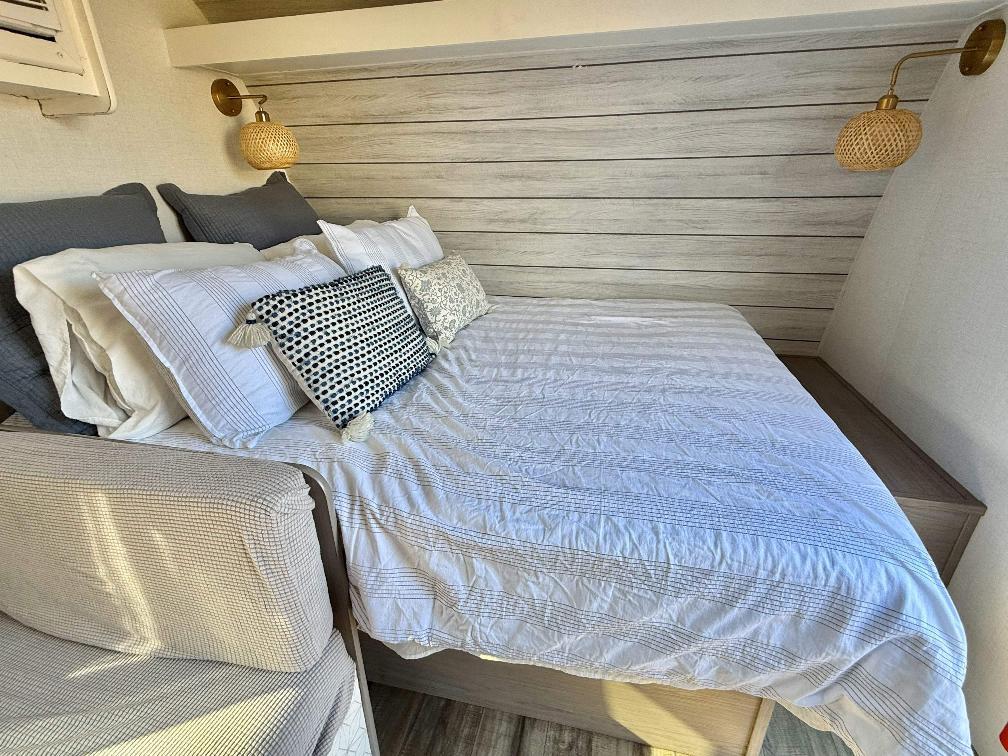 Cozy RV rental bedroom: bed with striped bedding, layered pillows, shiplap-style wall, and decorative woven sconce lights.. Dutchmen Coleman Lantern LT 2024
