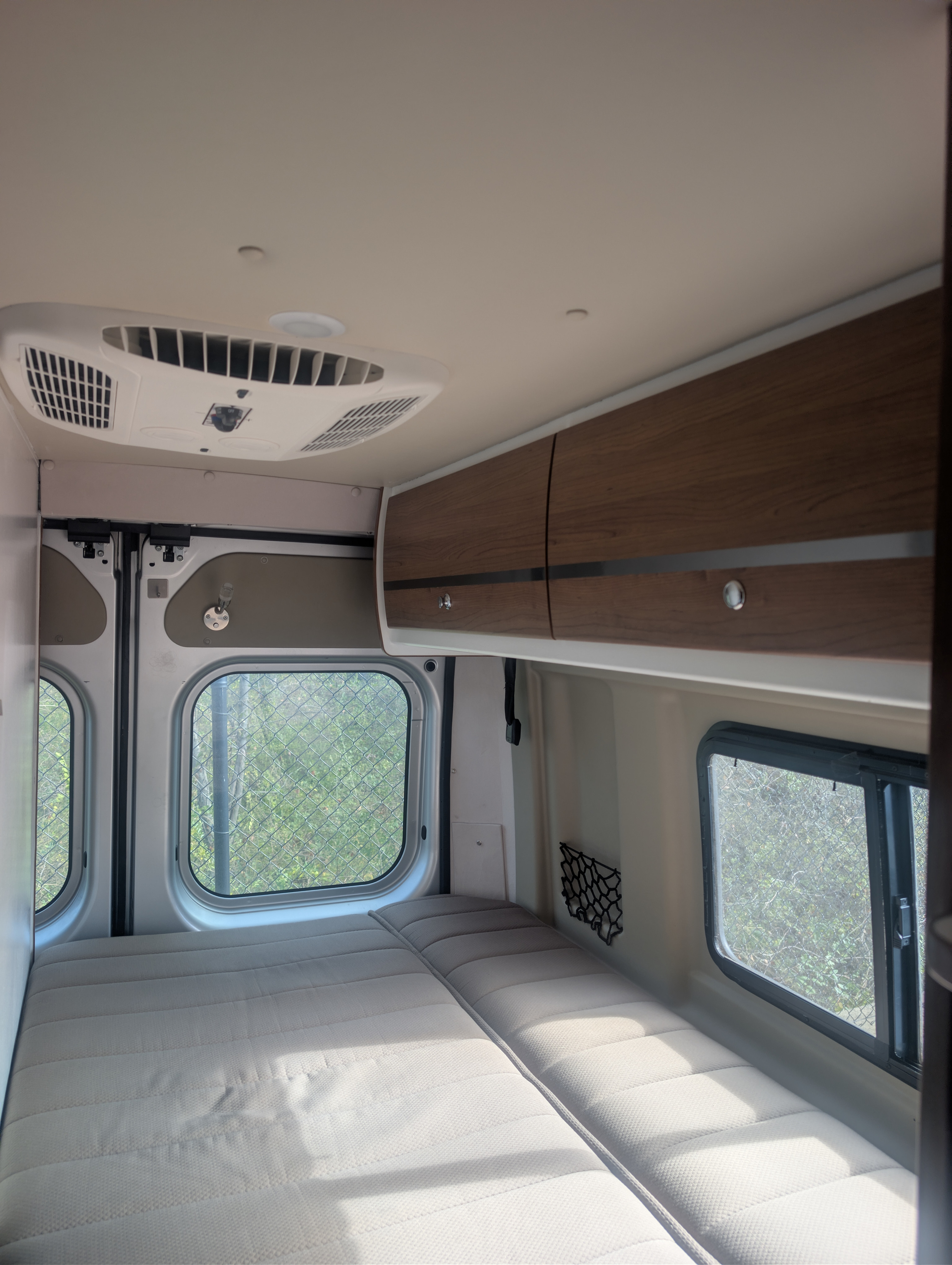 Cozy interior of an RV rental featuring a comfortable bed, windows, overhead cabinets, and an AC unit for your travels.. Winnebago Travato 2018