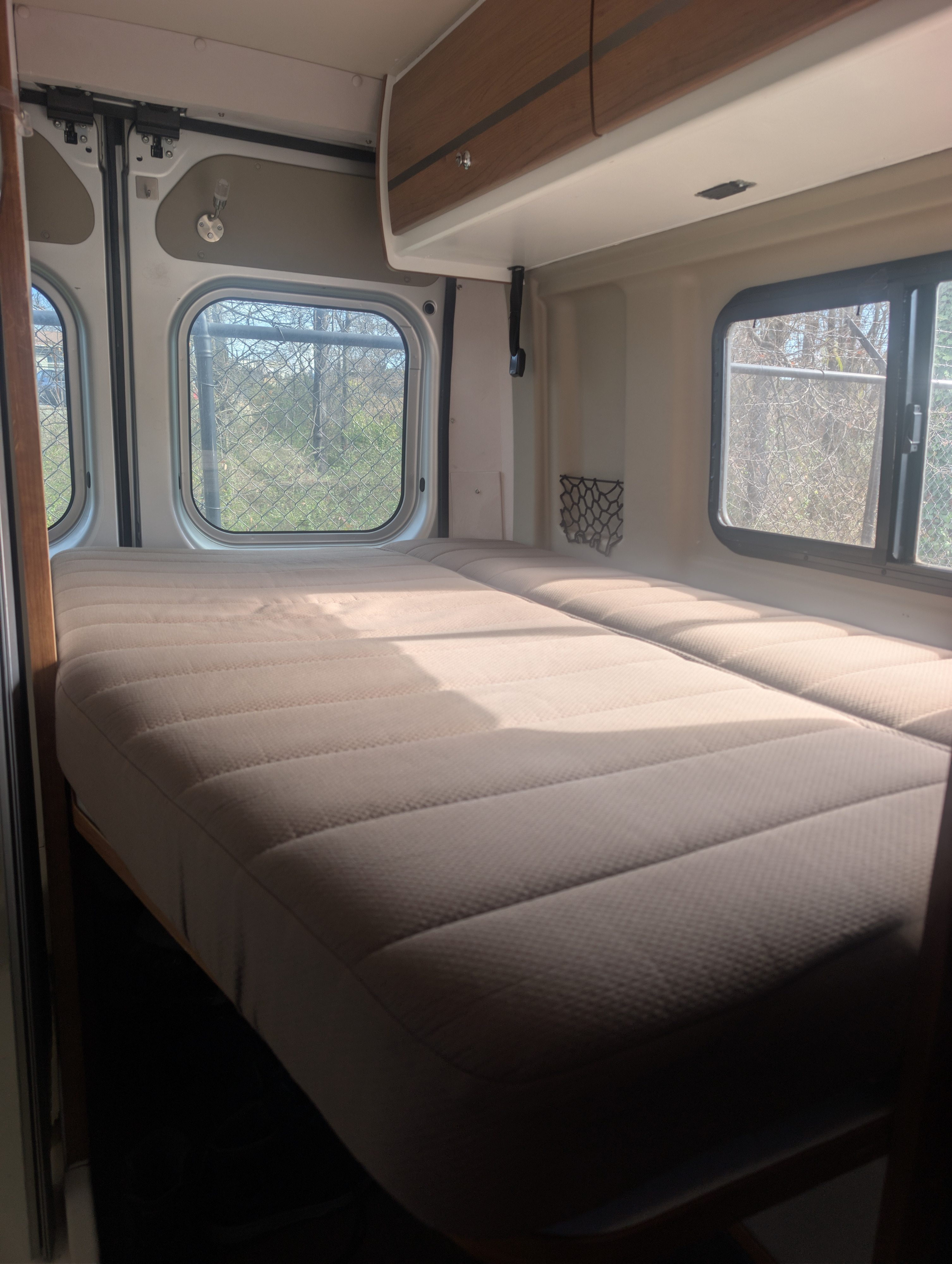 Bright rv rental interior with two beds, overhead cabinet, and side windows revealing trees and a fence outside, bathed in sunlight.. Winnebago Travato 2018