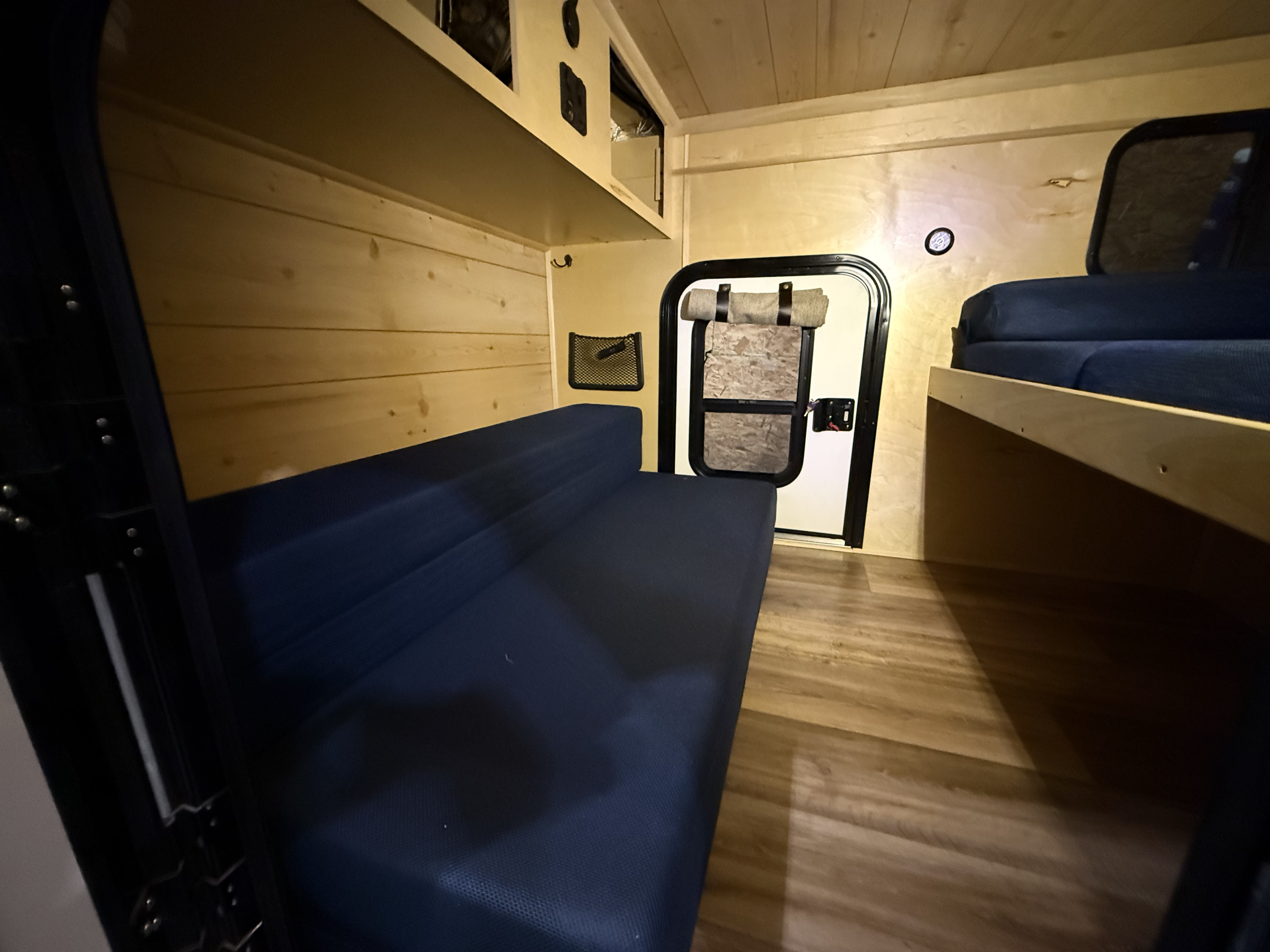 Cozy RV rental interior with warm wood paneling, featuring a blue bench bed, overhead storage, and a separate sleeping bunk.. CUSTOM ** GLAMPING - OFF GRID BUNK** 2021