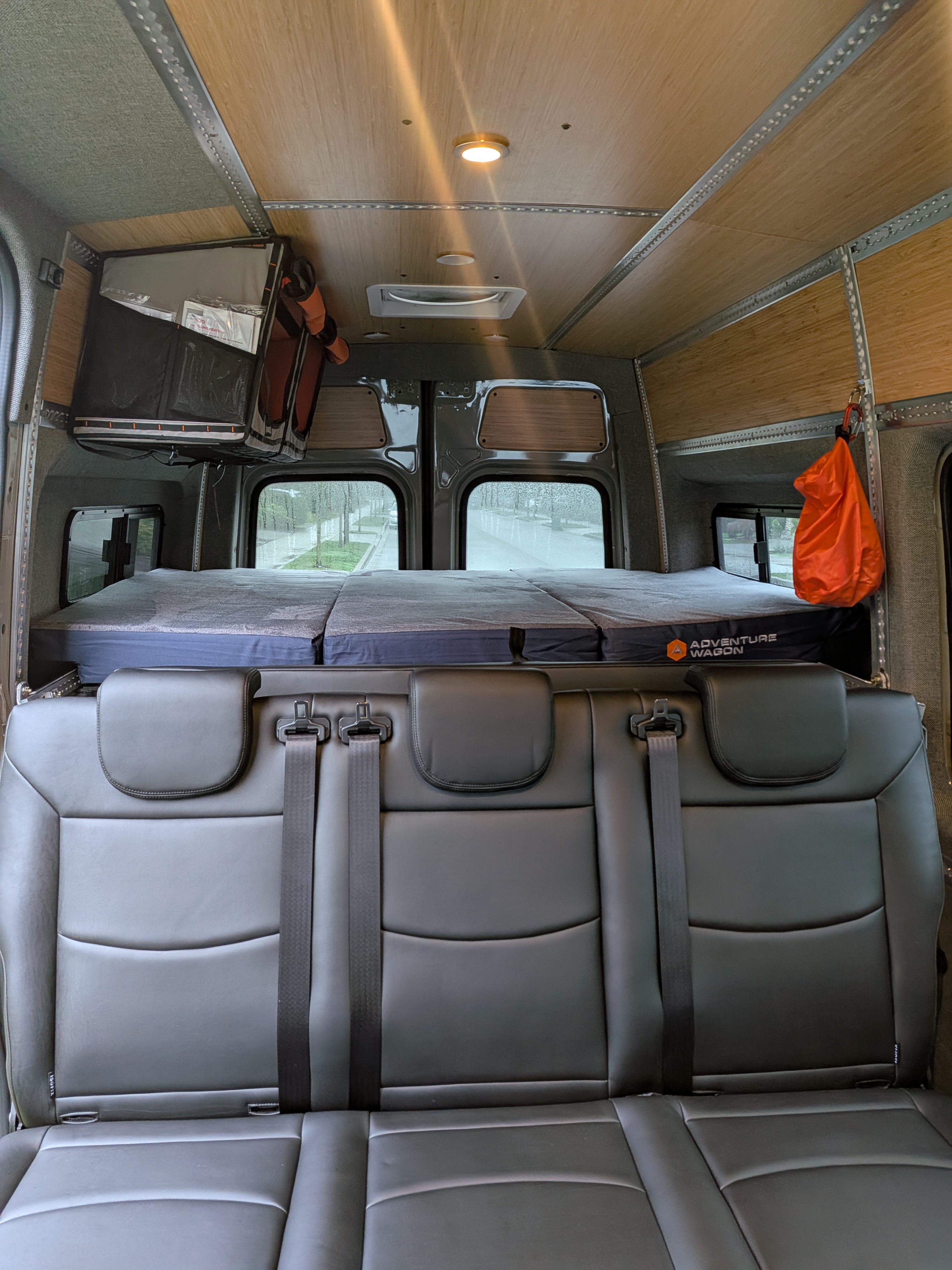 This rv rental interior features a 3-person black leather seat, custom bed with "Adventure Wagon" logo, wood-paneled ceiling, & windows showing a rainy day.. Mercedes-Benz Sprinter 2025
