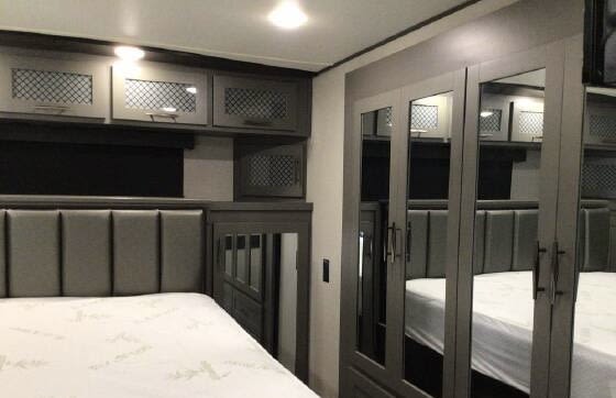 Relax in this RV rental bedroom featuring a comfy bed with a grey headboard, overhead cabinets, and spacious mirrored wardrobe.. Grand Design Momentum Toy Hauler 2021