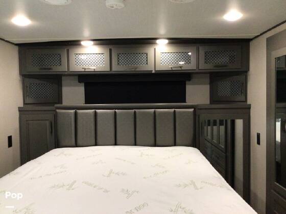 Cozy RV rental bedroom with a grey upholstered headboard, grid-patterned overhead cabinets, and built-in bedside storage.. Grand Design Momentum Toy Hauler 2021