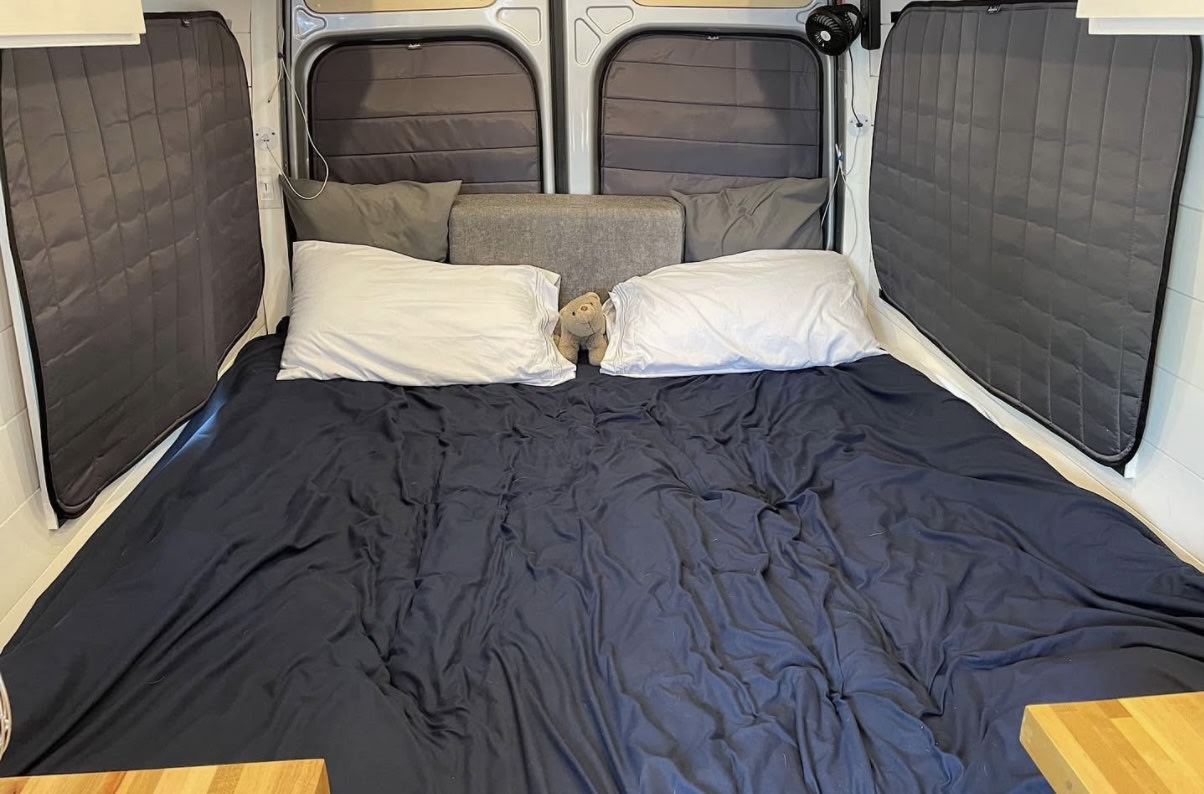Cozy RV rental bed in a camper van interior, featuring dark bedding, white pillows, and insulated windows.. Mercedes-Benz Sprinter 2020
