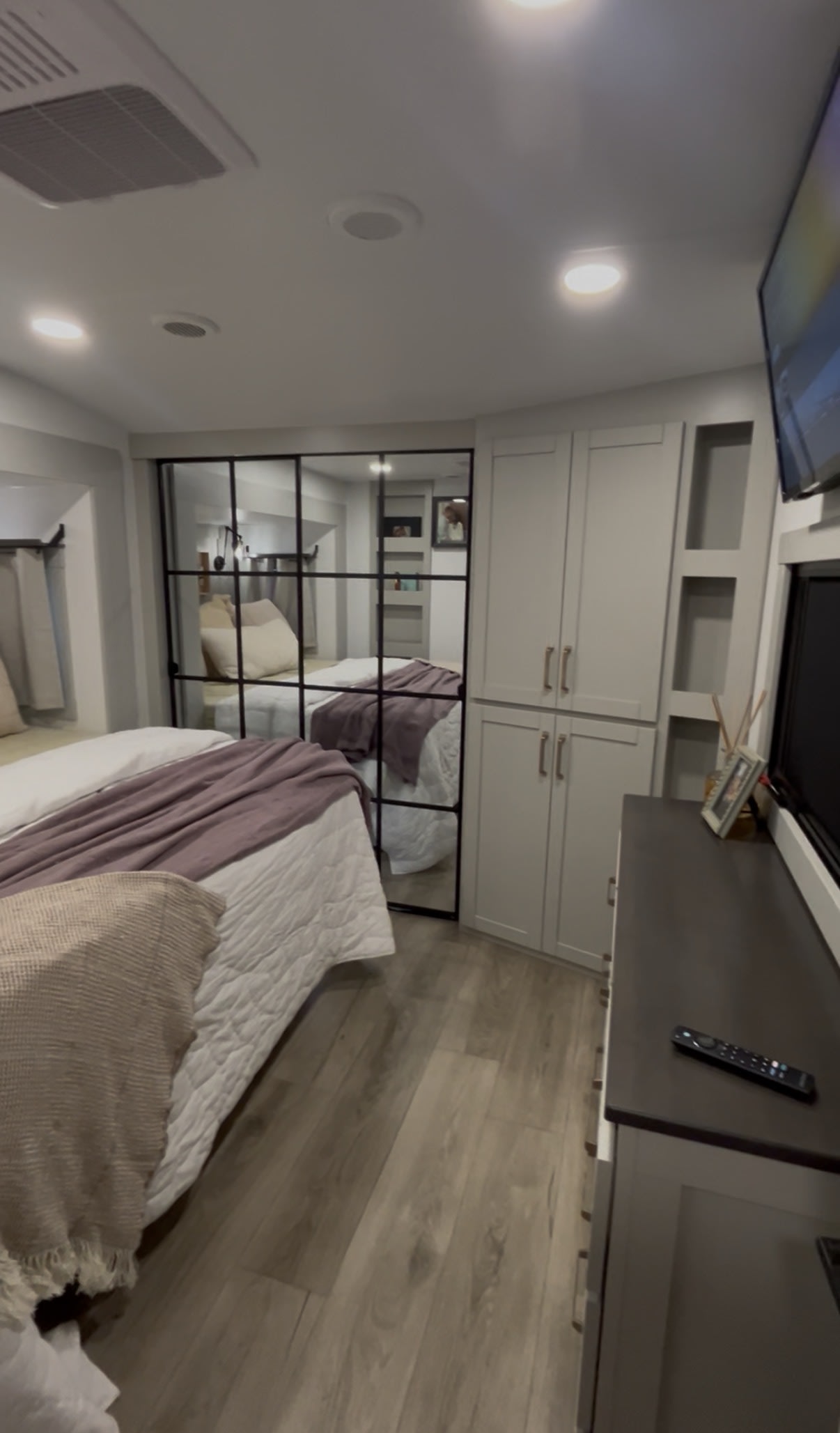 Modern RV rental bedroom: bed with white and purple bedding, black-framed mirror, built-in cabinets, TV, and wood-look floor.. Brinkley RV Model Z 2025