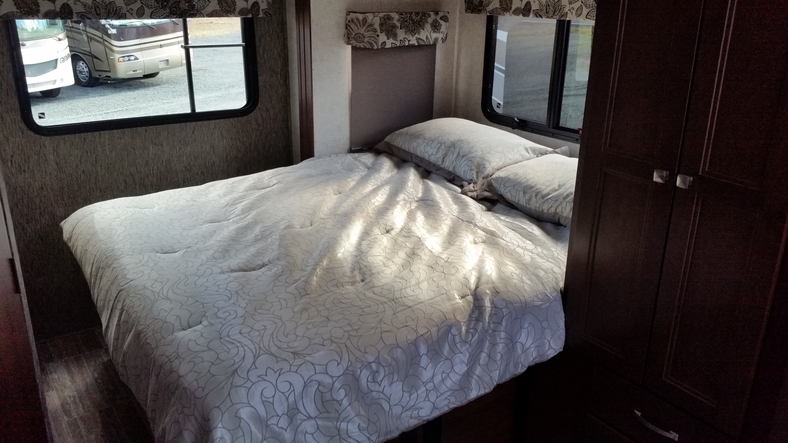 Bright RV rental bedroom features a comfy bed with patterned bedding, pillows, a window view of other RVs, and a dark wardrobe.. Coachmen 2401W 2018