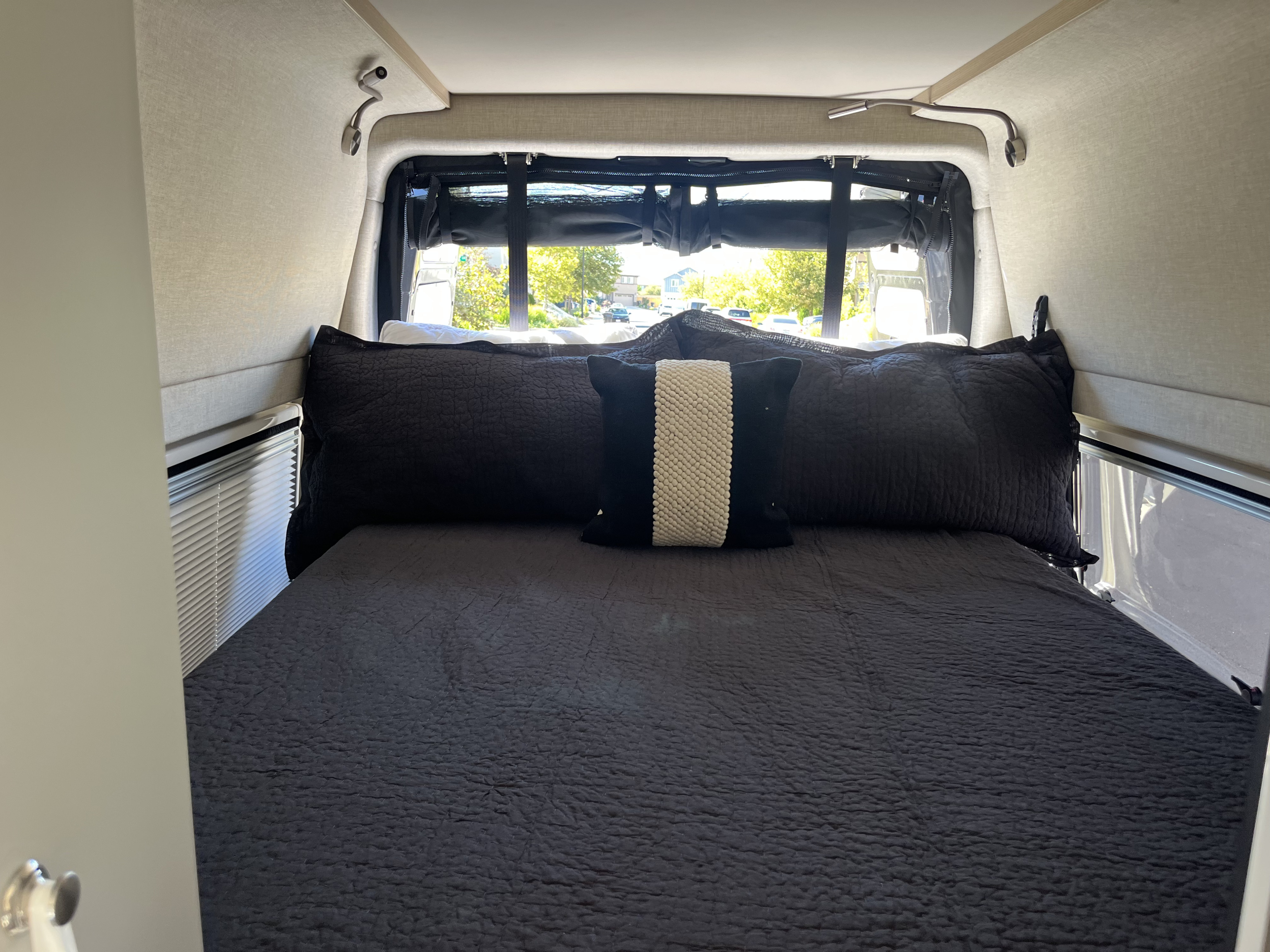 Cozy RV rental bedroom with dark quilted bedding, accent pillow, reading lights, and an outside view from the rear window.. Noovo Plus LB 2025
