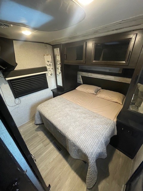 Inside an RV rental bedroom: cozy bed with light bedding, overhead cabinets, TV, and dark wood accents.. Jayco Jay Feather 2021