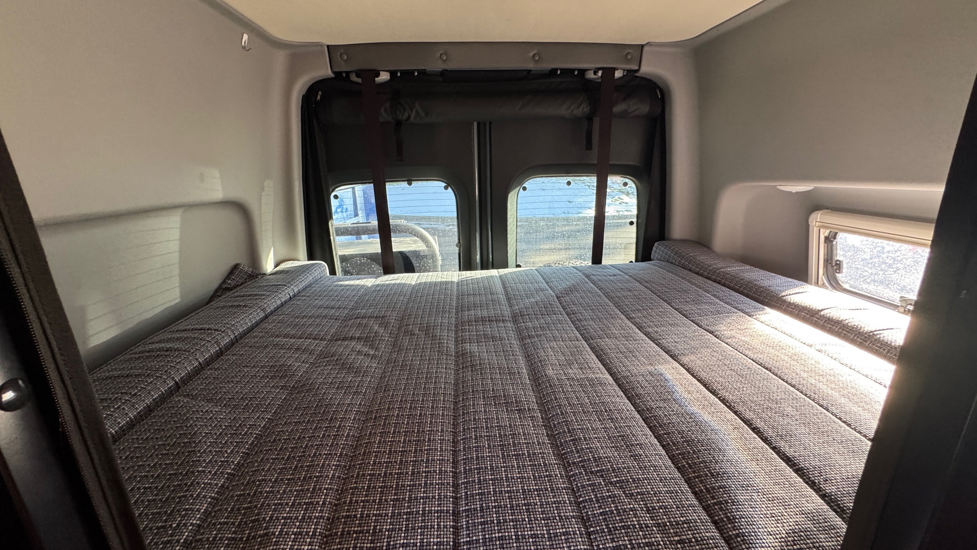 Inside an RV rental: a comfortable, patterned bed ready for rest, with bright windows revealing the exterior view.. Winnebago Revel 2023