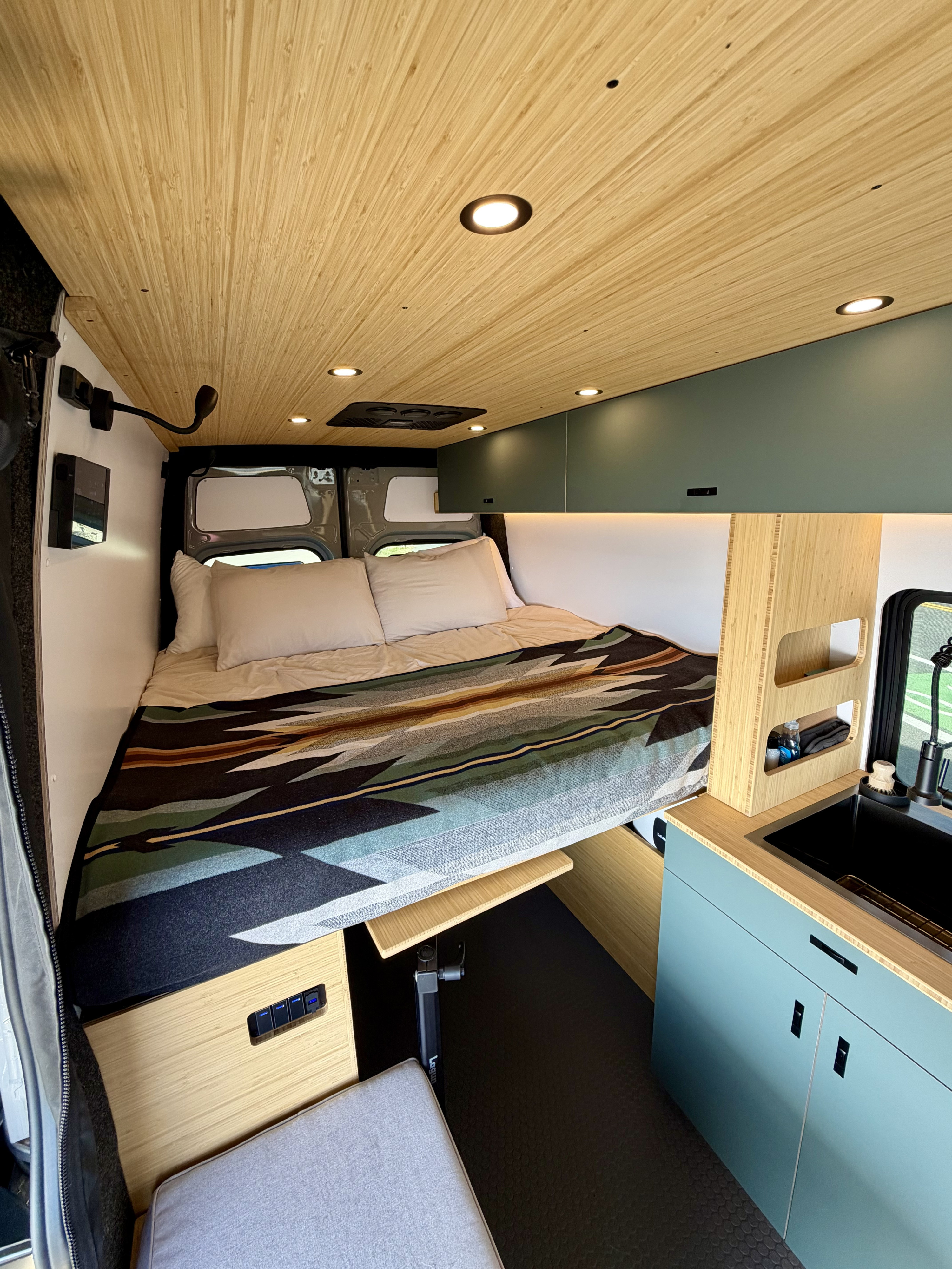 RV rental's modern interior: bamboo ceiling, cozy bed with patterned blanket, kitchenette & smart storage.. Mercedes-Benz Sprinter 2025