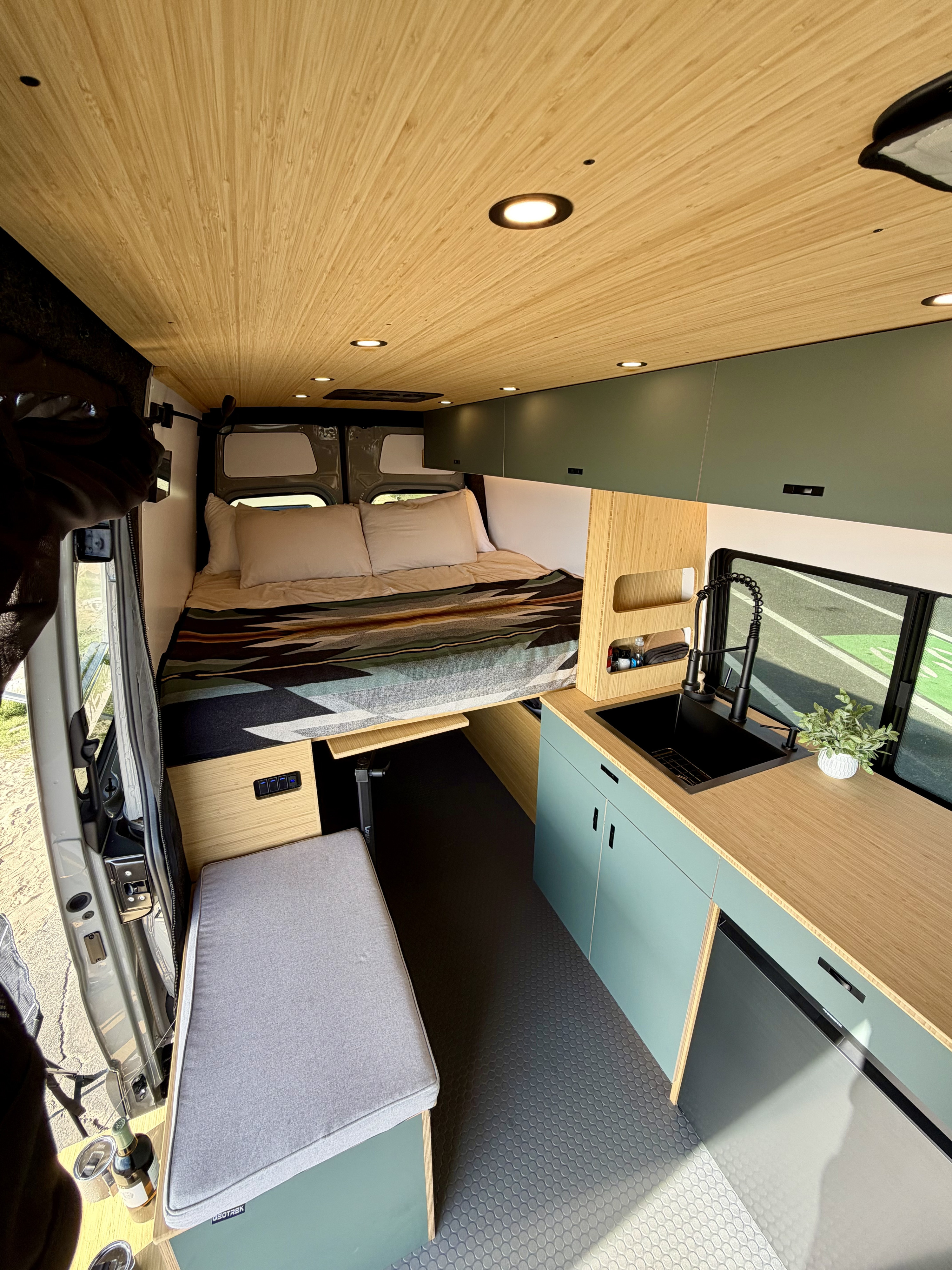Modern rv rental camper van interior: wood ceiling, cozy bed, sleek kitchen with black sink, light green cabinets, and seating.. Mercedes-Benz Sprinter 2025