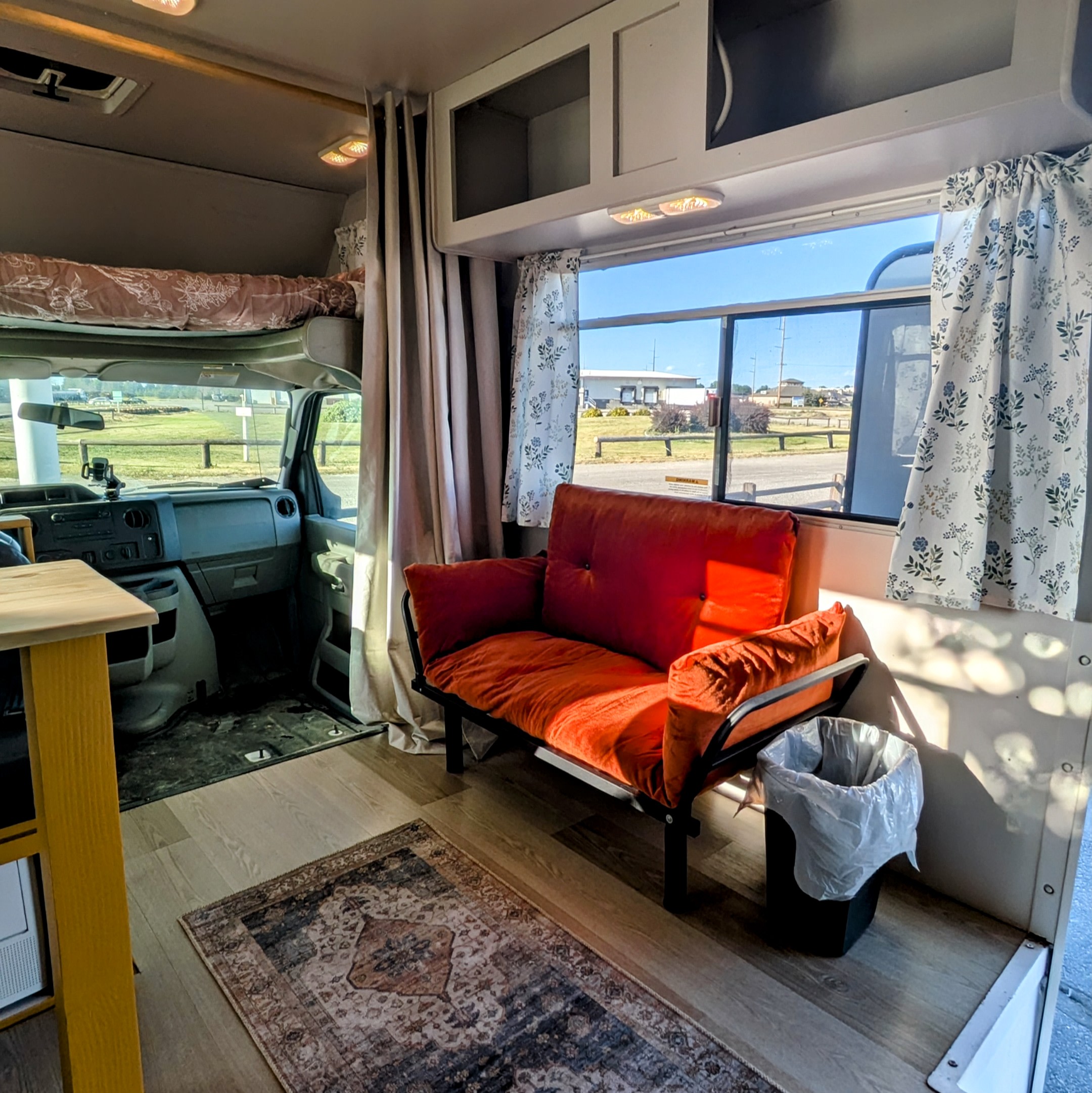 Cozy RV rental interior with an orange futon, window views, and an overhead bed, perfect for your road trip comfort.. Thor Majestic 2019