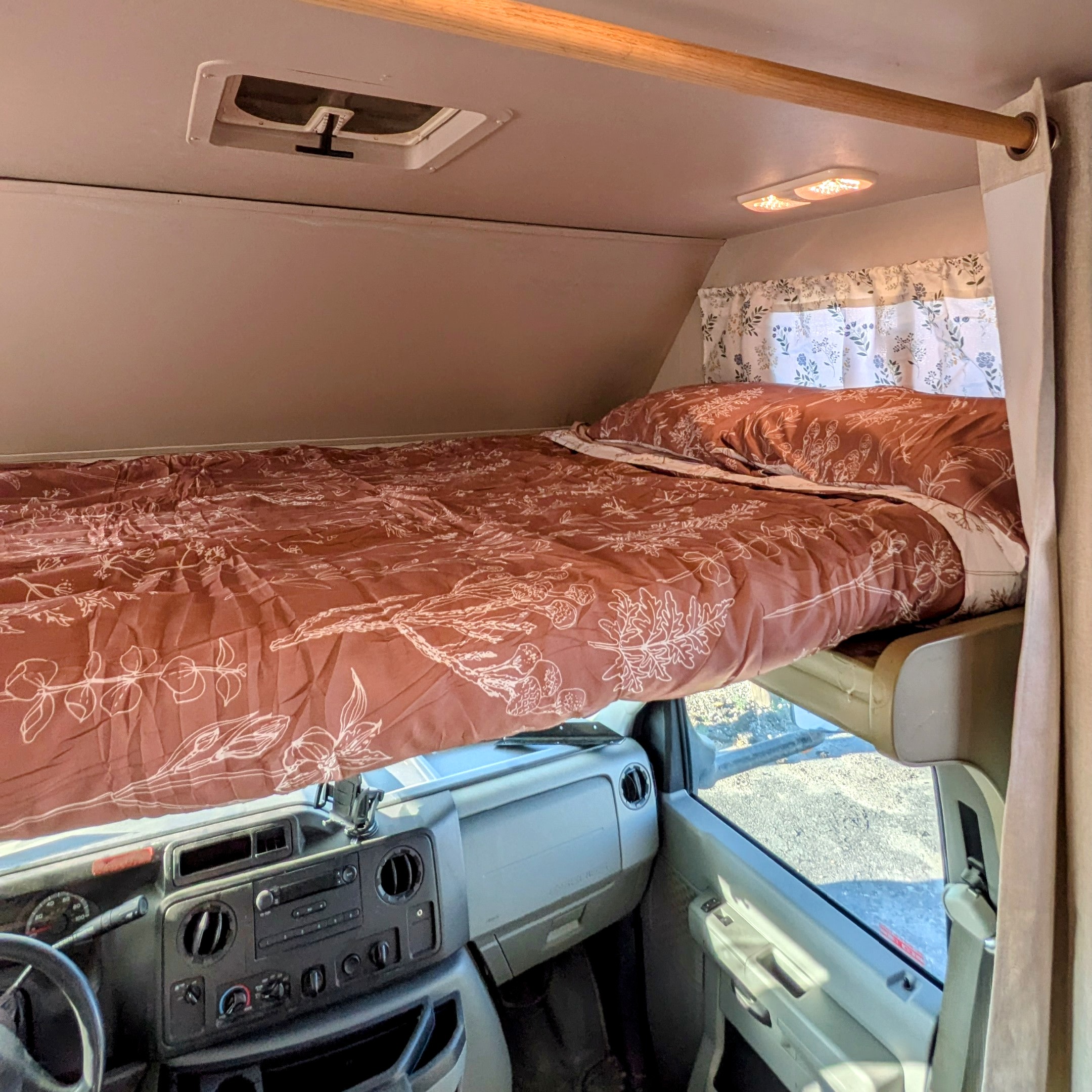 Inside an **rv rental**: a comfortable cab-over bunk bed with a floral duvet, overhead lights, and the driver's dashboard below.. Thor Majestic 2019