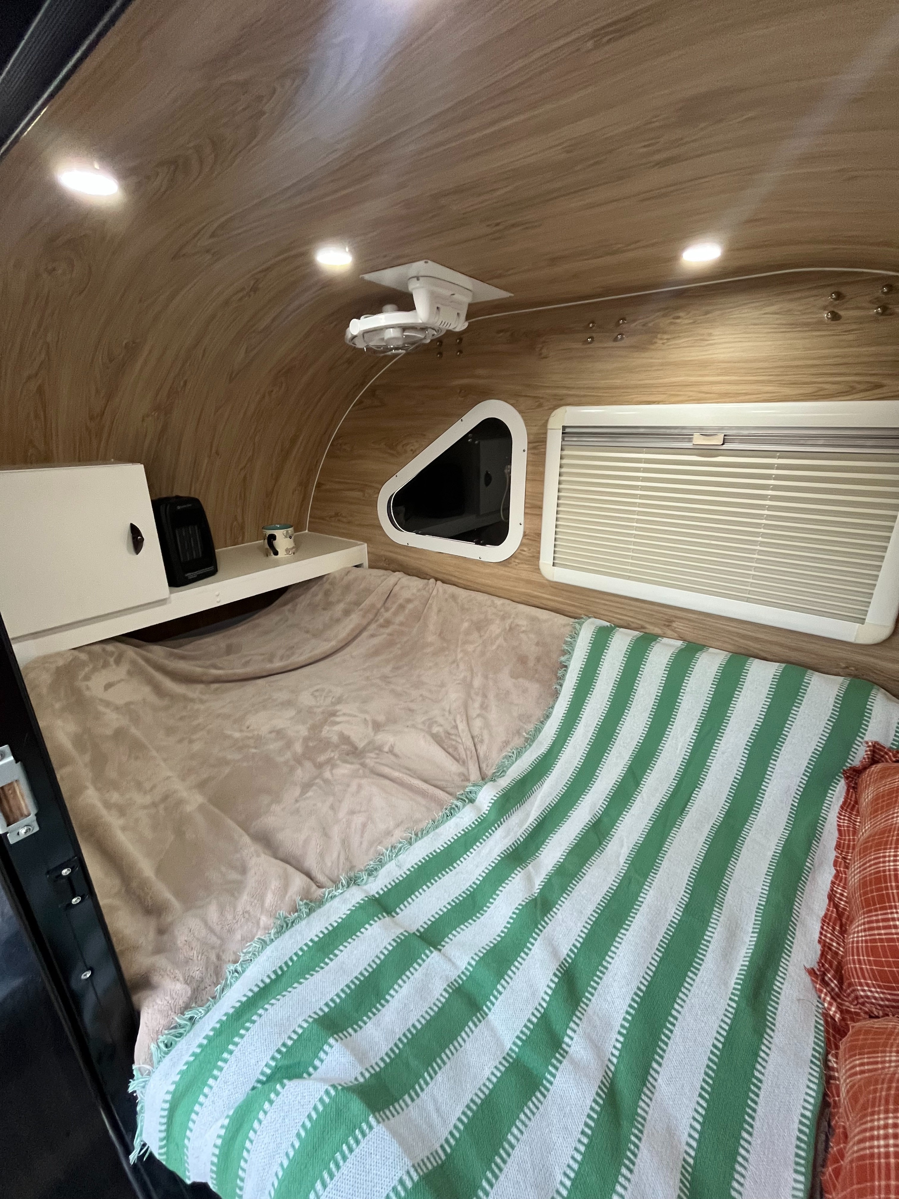 Cozy RV rental interior features a comfortable bed with a green blanket, wood-paneled ceiling, shelf, and windows.. Ecocamper Vehicle Co. Eco camper 2023