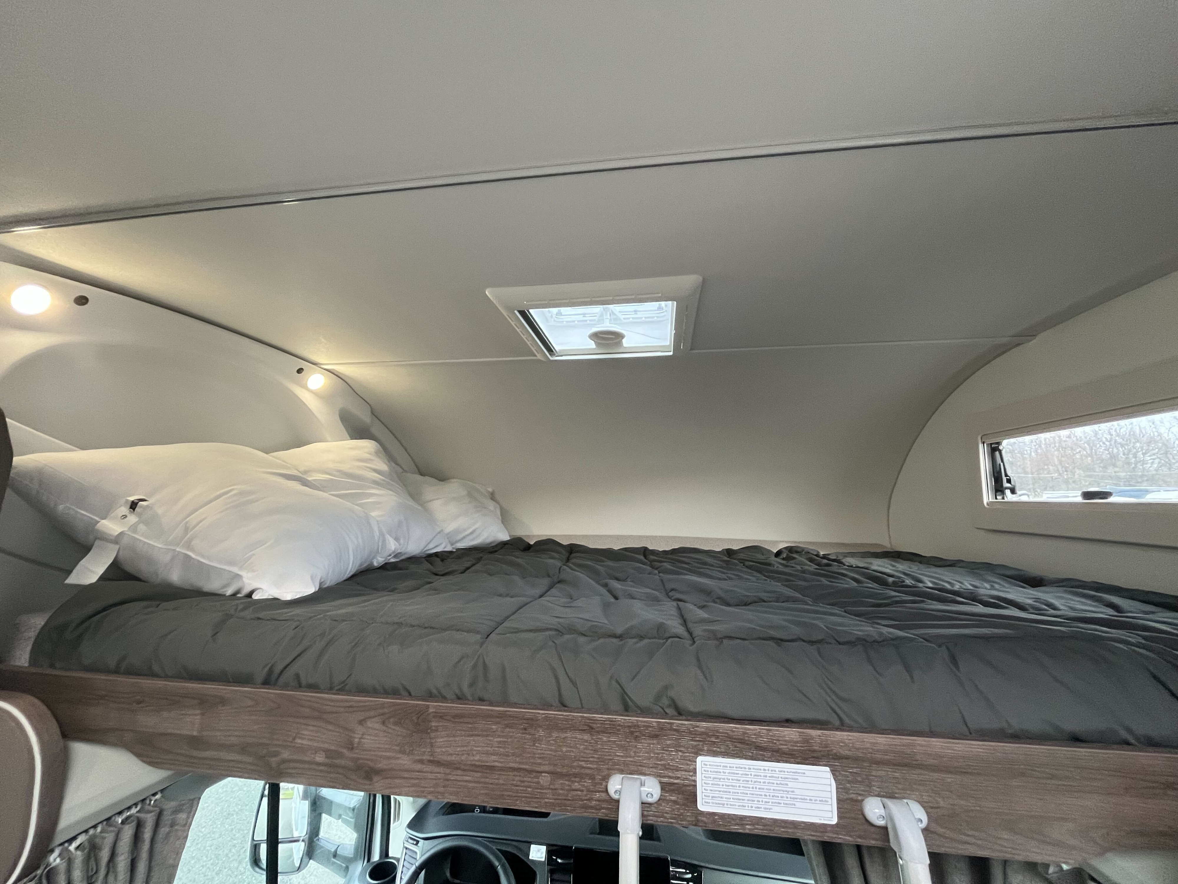 This RV rental's overcab bed features a dark grey comforter, white pillows, built-in lights, and a ceiling skylight, ideal for a cozy rest.. Benimar Sport 324 UP 2025