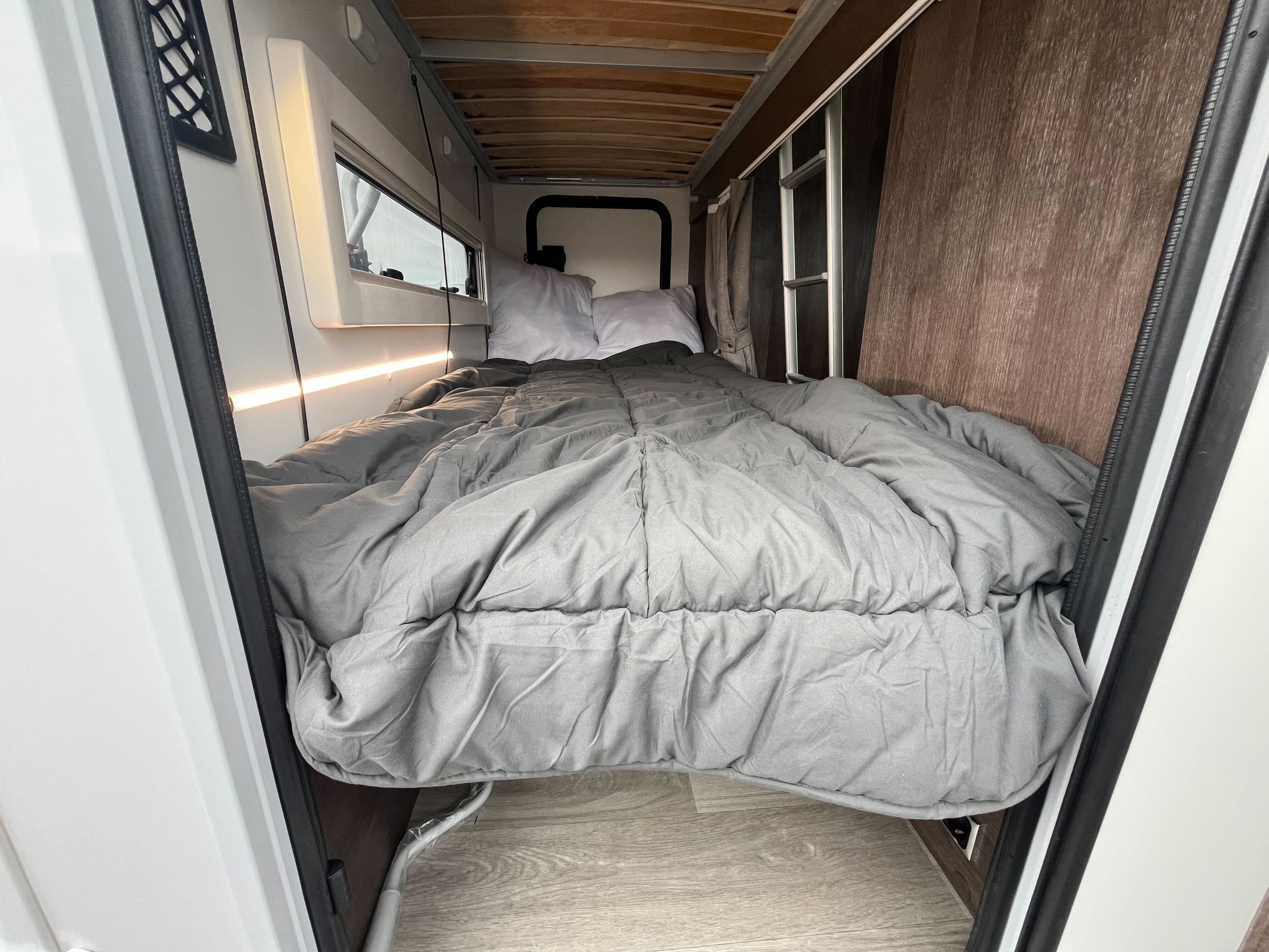 Inside an RV rental, a comfy bed with grey comforter and pillows awaits. Features a window, LED lighting, and a bunk ladder.. Benimar Sport 324 UP 2025