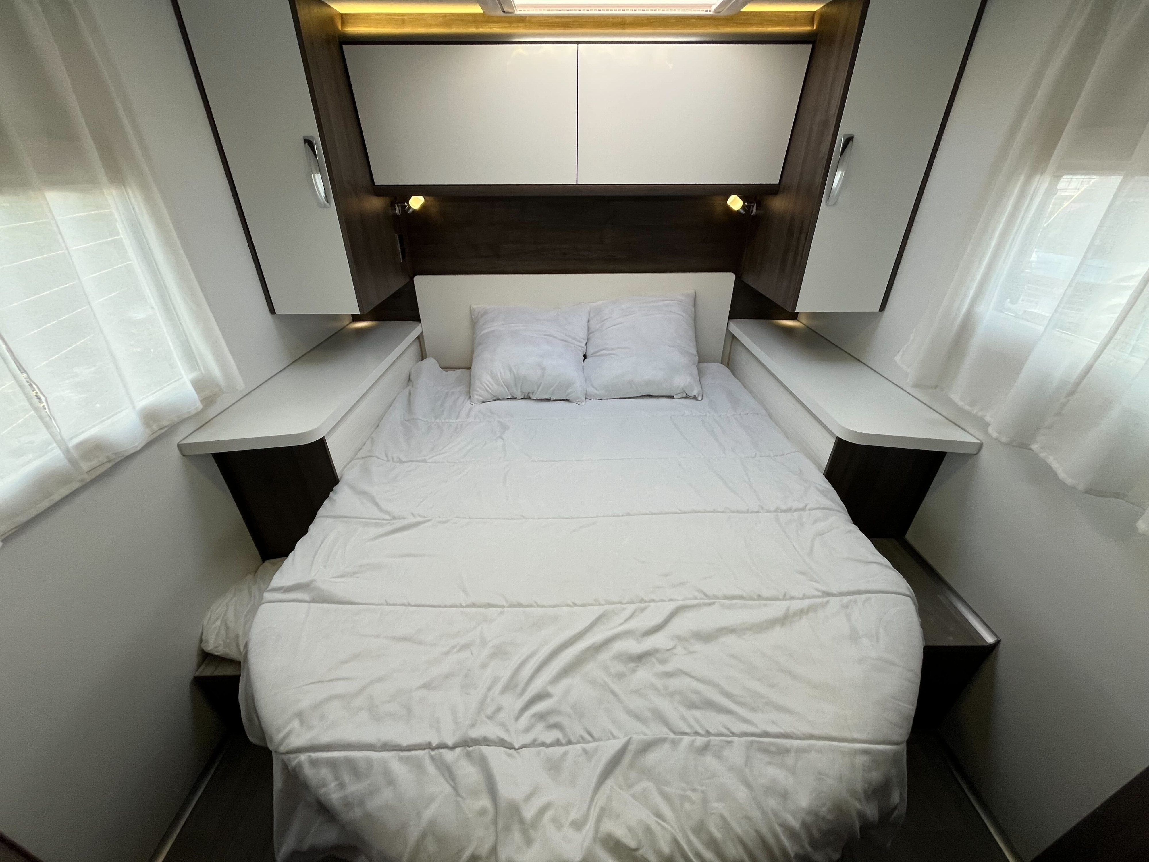 Cozy RV rental bedroom with white bed, overhead storage, and reading lights, offering a comfortable sleeping area.. BENIMAR T 497 TESSORO 2025