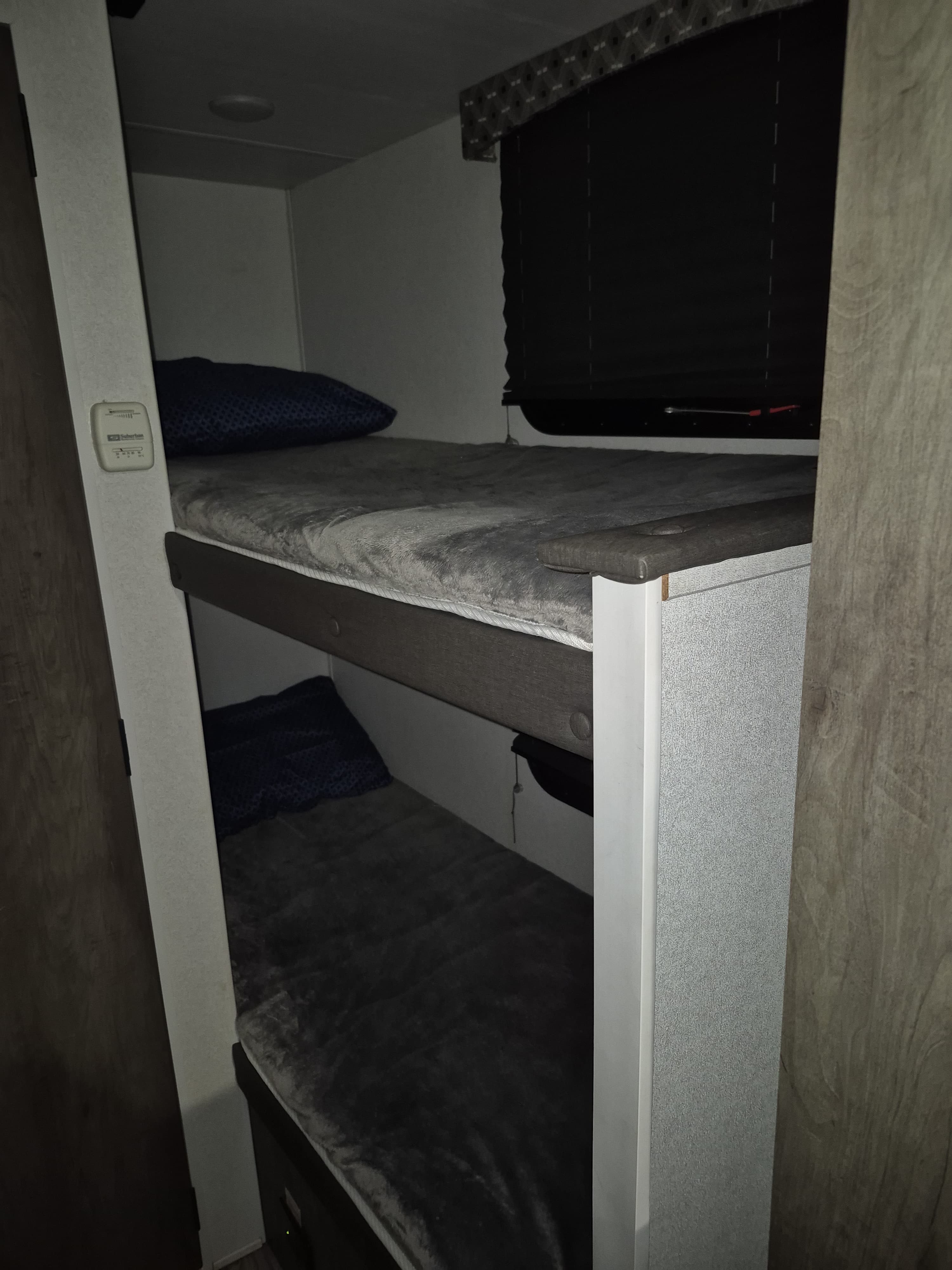 Interior of an RV rental showing two bunk beds with soft grey bedding, a wall thermostat, and window blinds, set in dim lighting.. Forest River Sonoma 2020