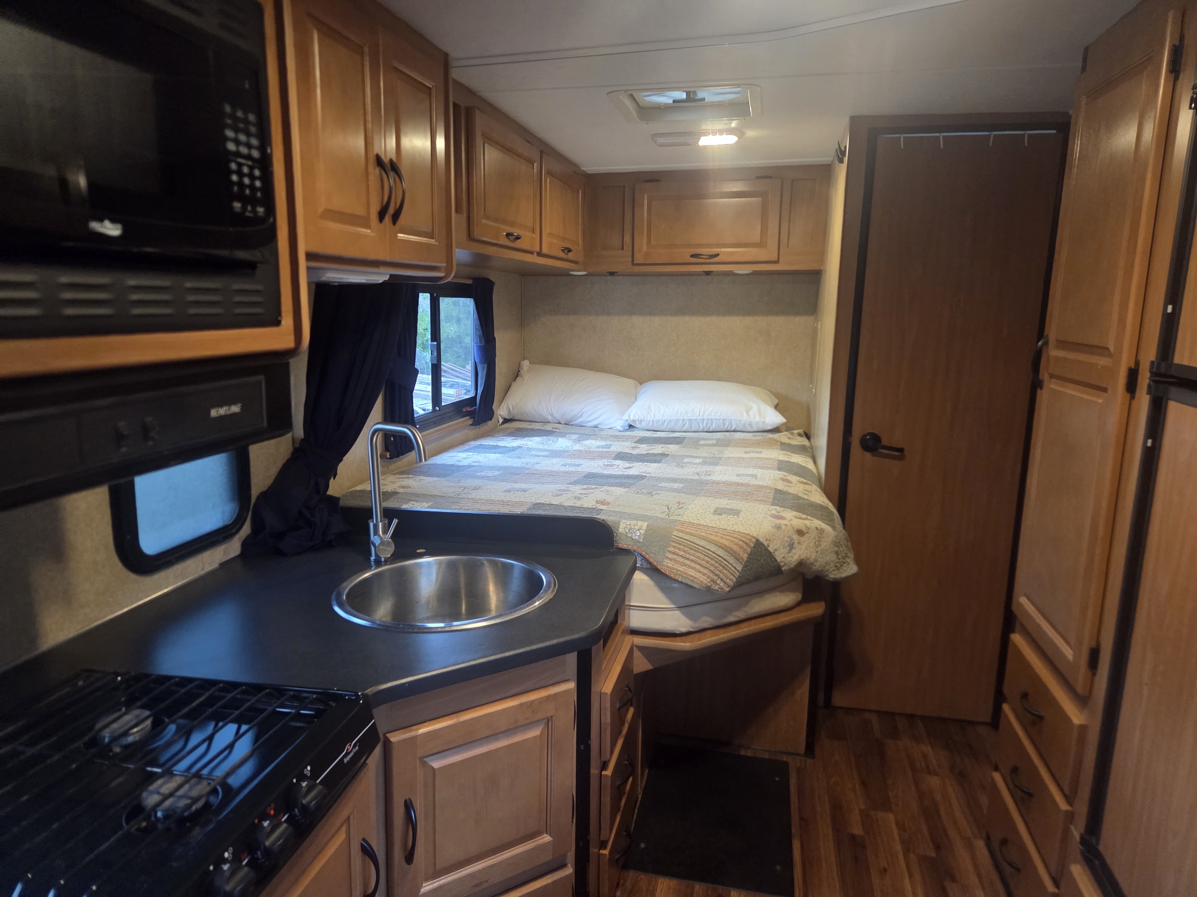 RV rental interior featuring a compact kitchenette with microwave, sink, and stove, plus a cozy bed and wooden cabinets.. Thor Majestic 2017
