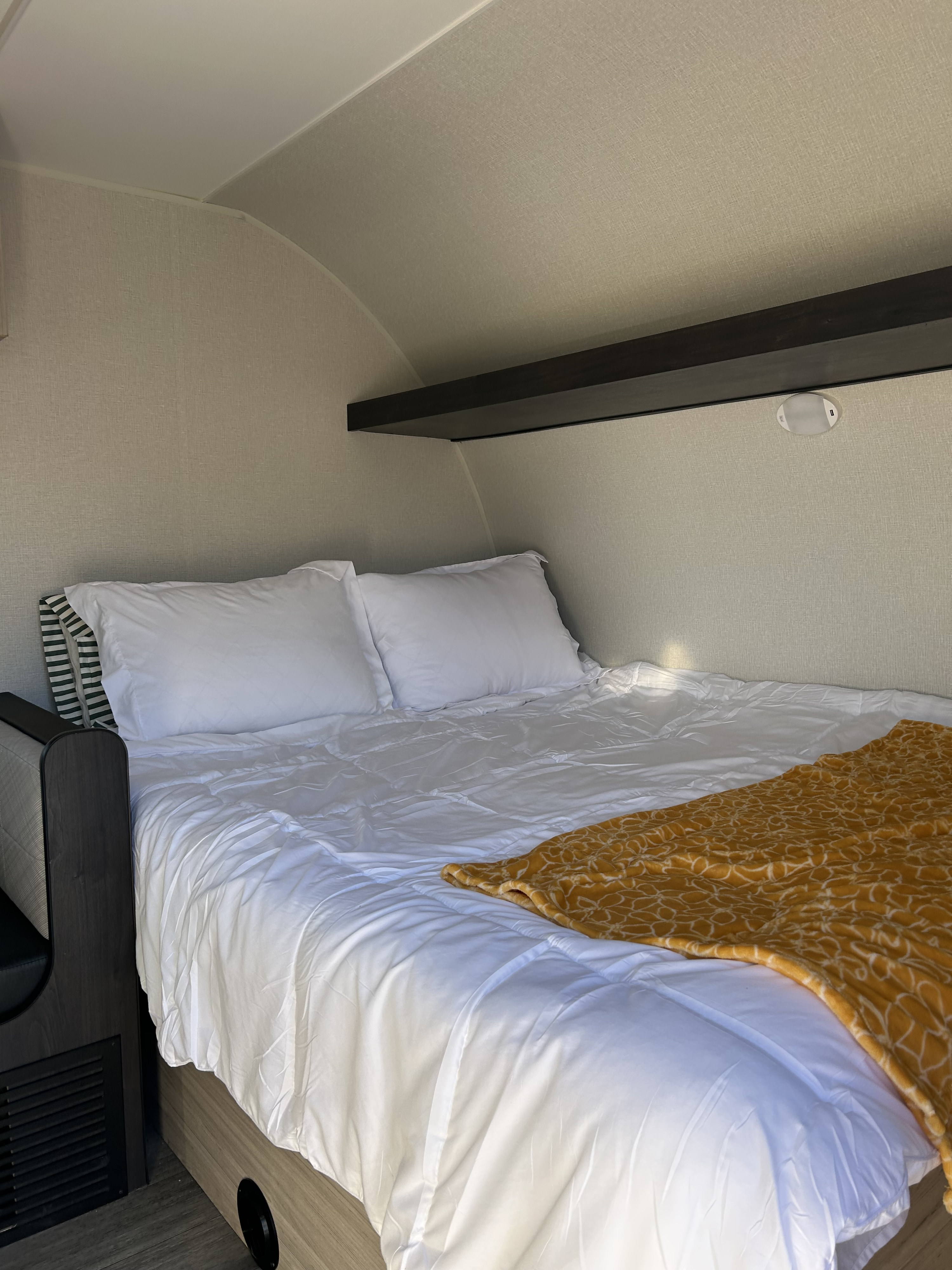 This cozy RV rental bedroom has a bed with crisp white bedding, two pillows, and a cheerful yellow blanket, beneath a curved ceiling and storage shelf.. Heartland Mallard 2025