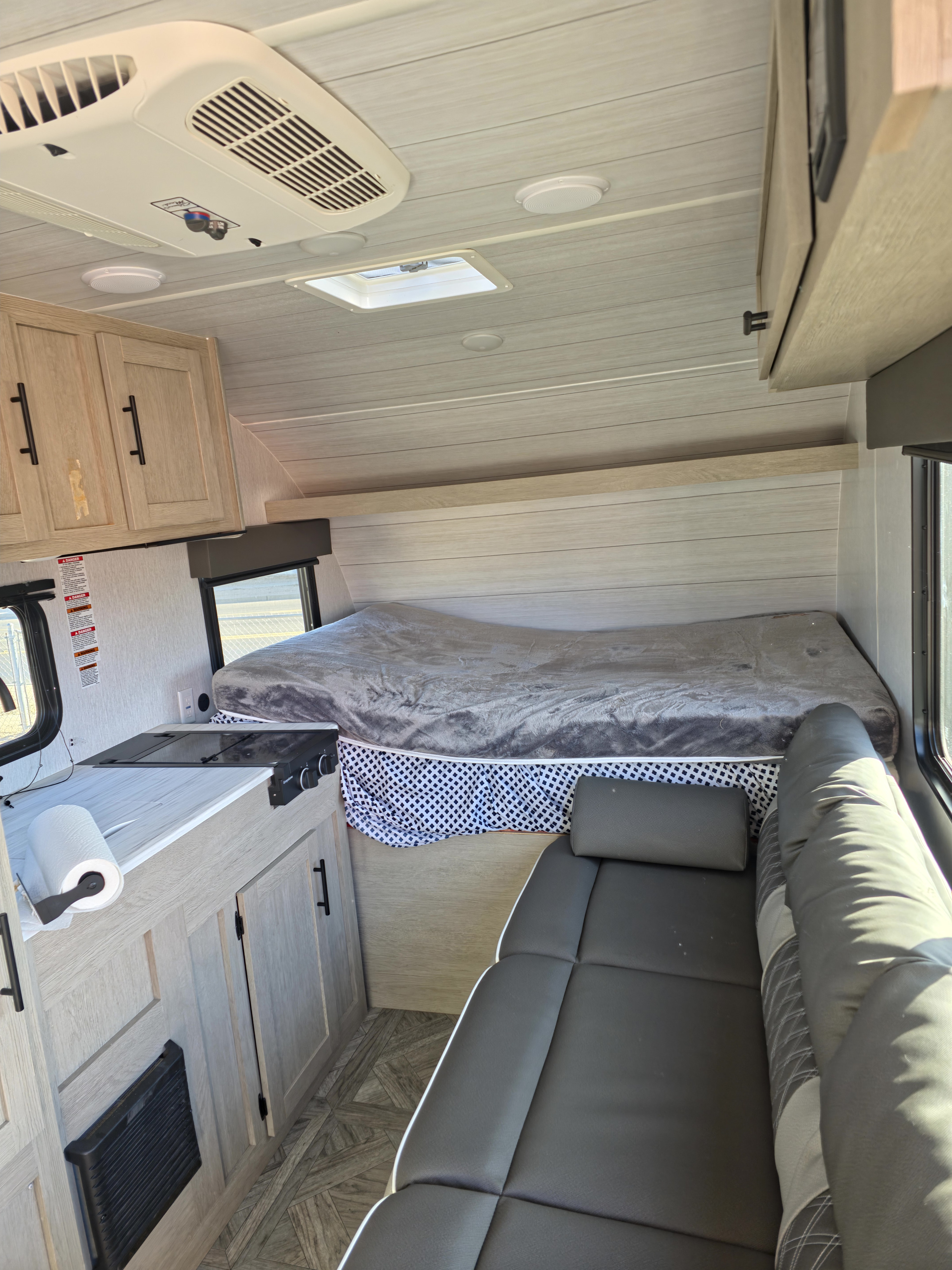Modern RV rental interior: kitchenette, raised bed with grey blanket, and dark grey cushioned seating. Bright, compact camper space.. Forest River Evo G.O.A.T 2024