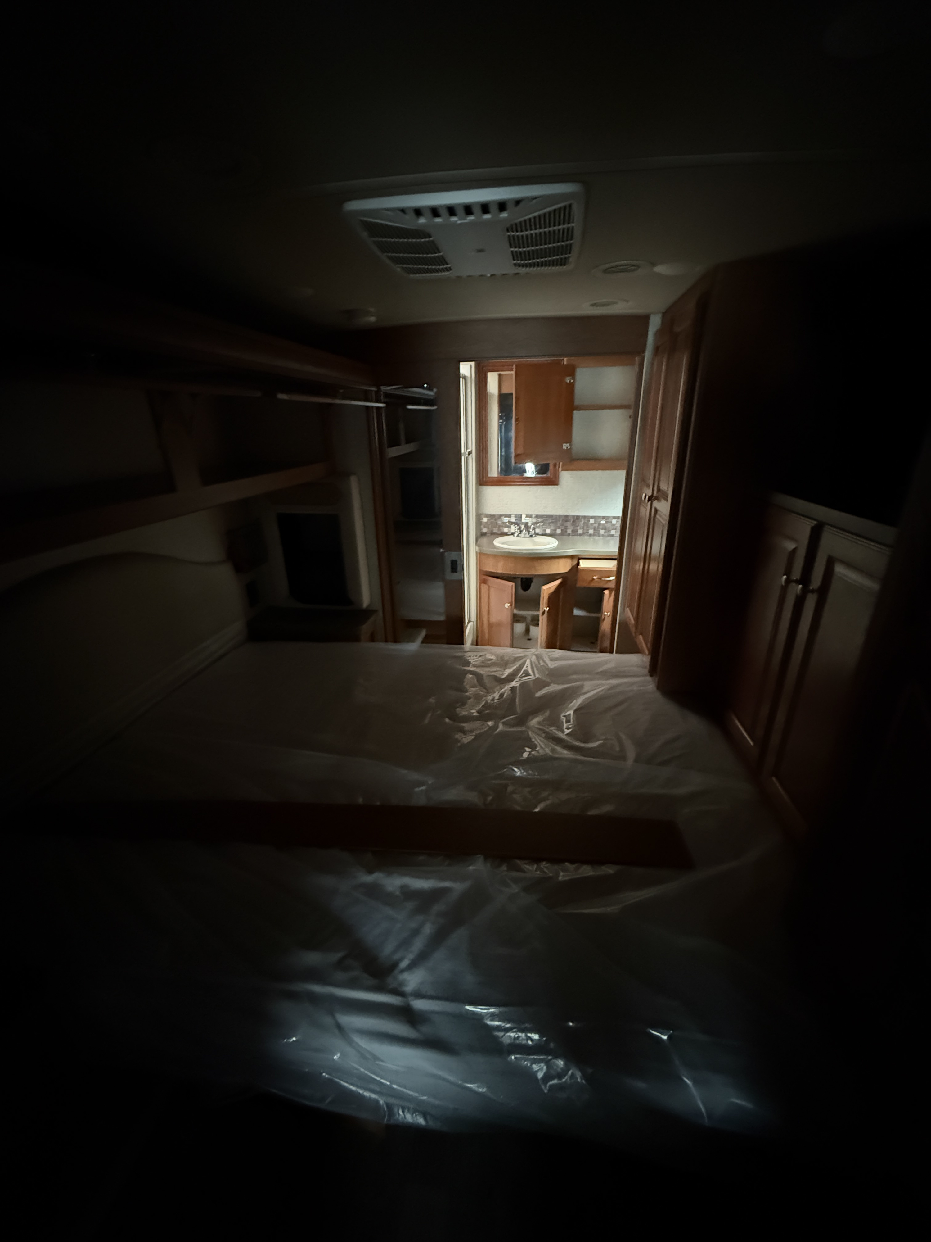 Dimly lit RV rental bedroom and bathroom, with a plastic-covered bed and wooden cabinets.. Winnebago Sunstar LX 2017