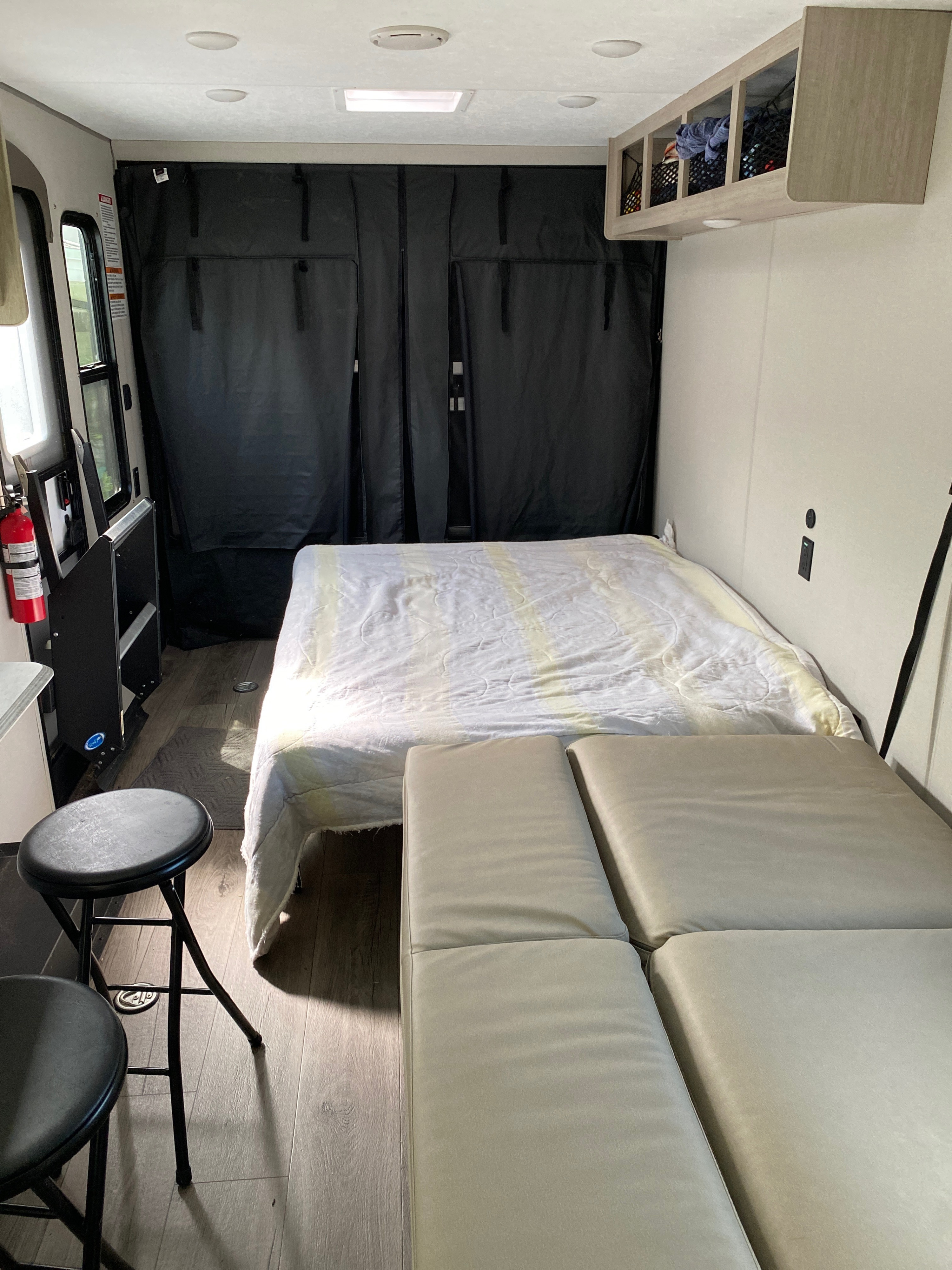 Inside an RV rental, showing a bed with white bedding, modular seating, overhead storage, and two stools on wood-look flooring.. Forest River Catalina 2024