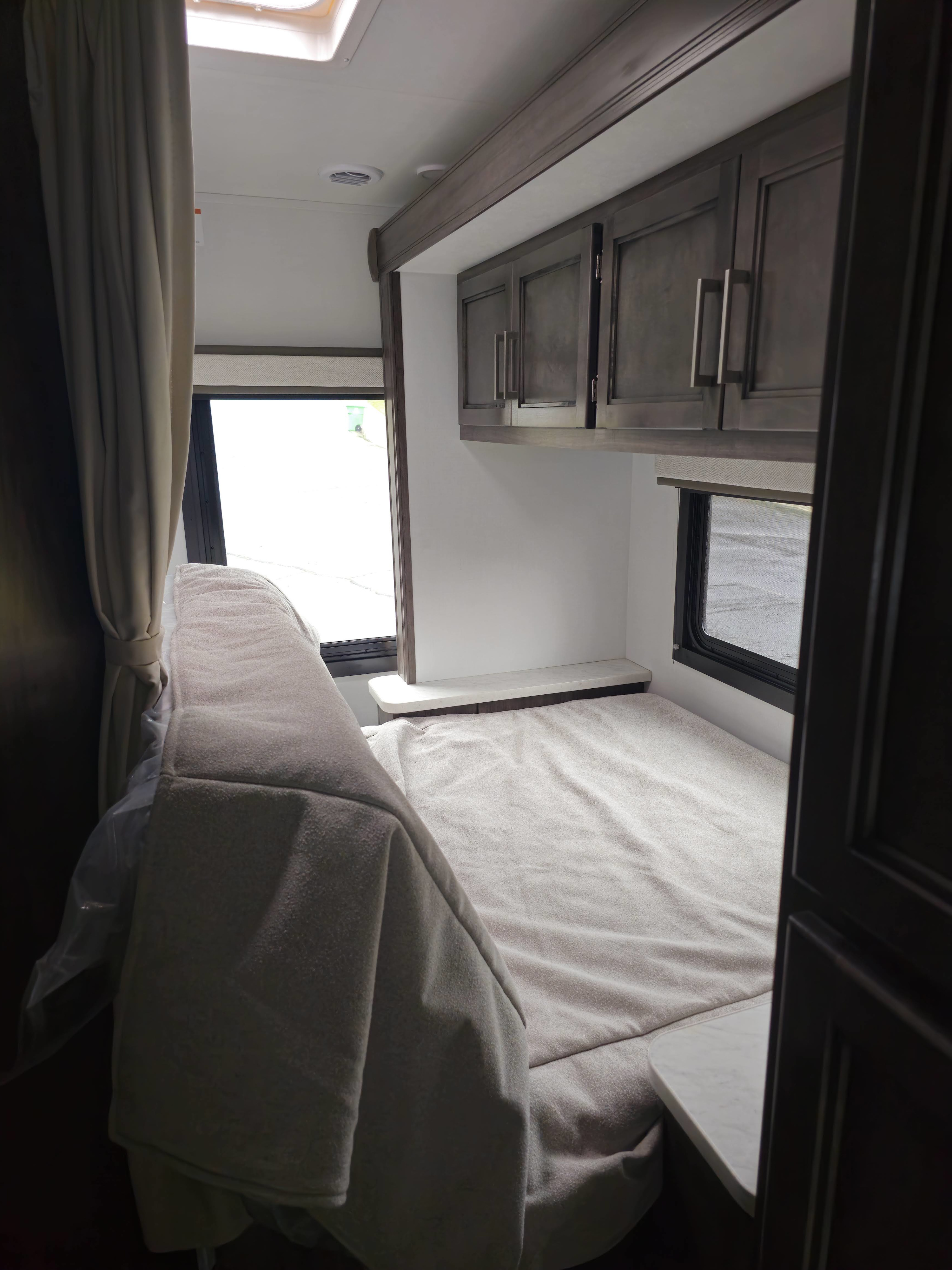 Inside this RV rental's bedroom: a tidy bed, dark overhead cabinets, a bright window, and light curtains.. Leisure Travel Vans Odyssey 22C 2026