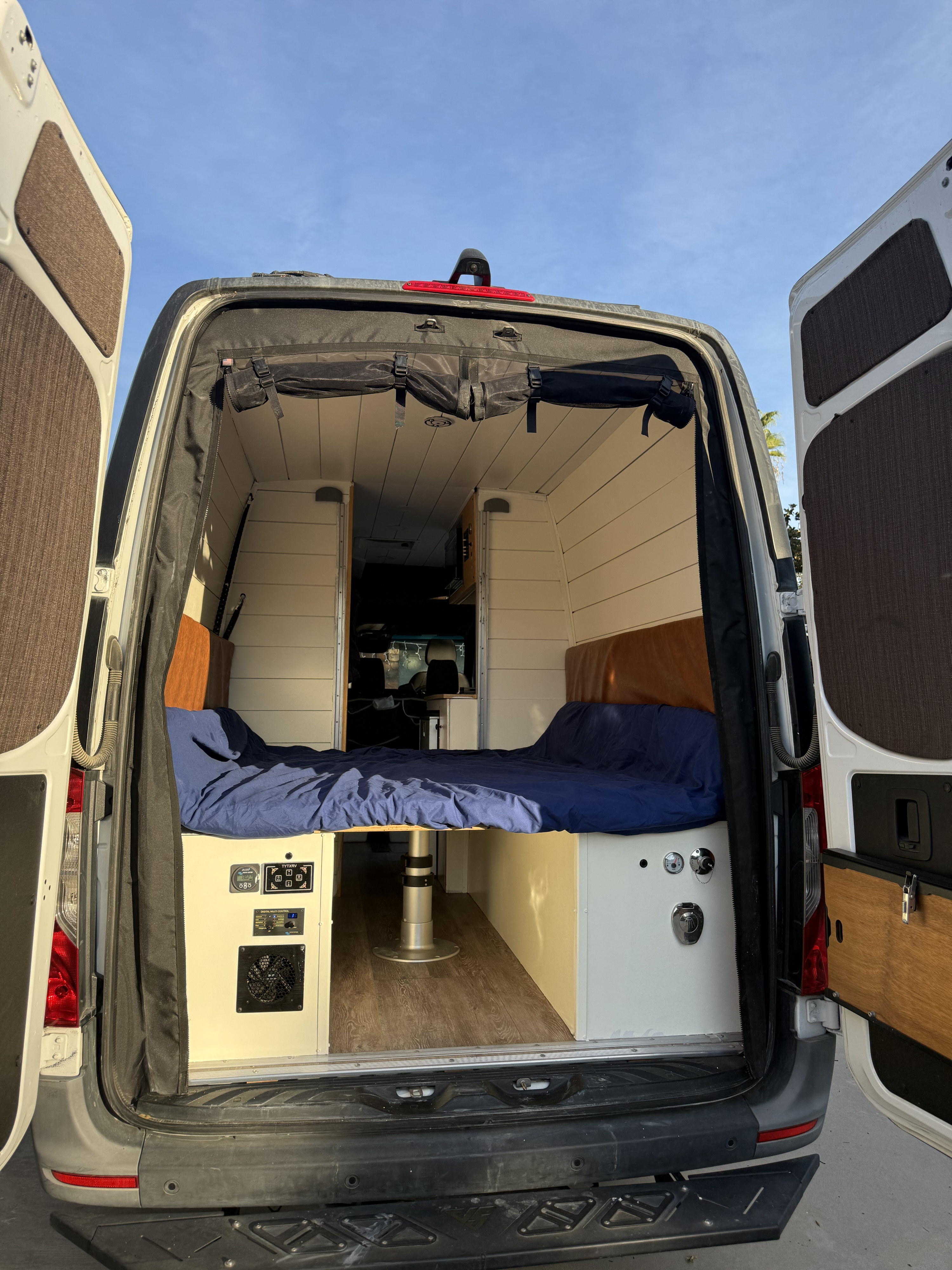 Step inside this cozy RV rental camper van! Features include a custom bed, control panel, and open rear doors.. Mercedez-Benz Sprinter 2021