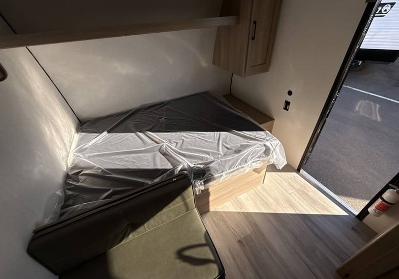 Inside a compact **rv rental** bedroom area: a plastic-wrapped bed, overhead cabinet, and part of a dinette. Light wood accents.. Keystone Coleman 2026