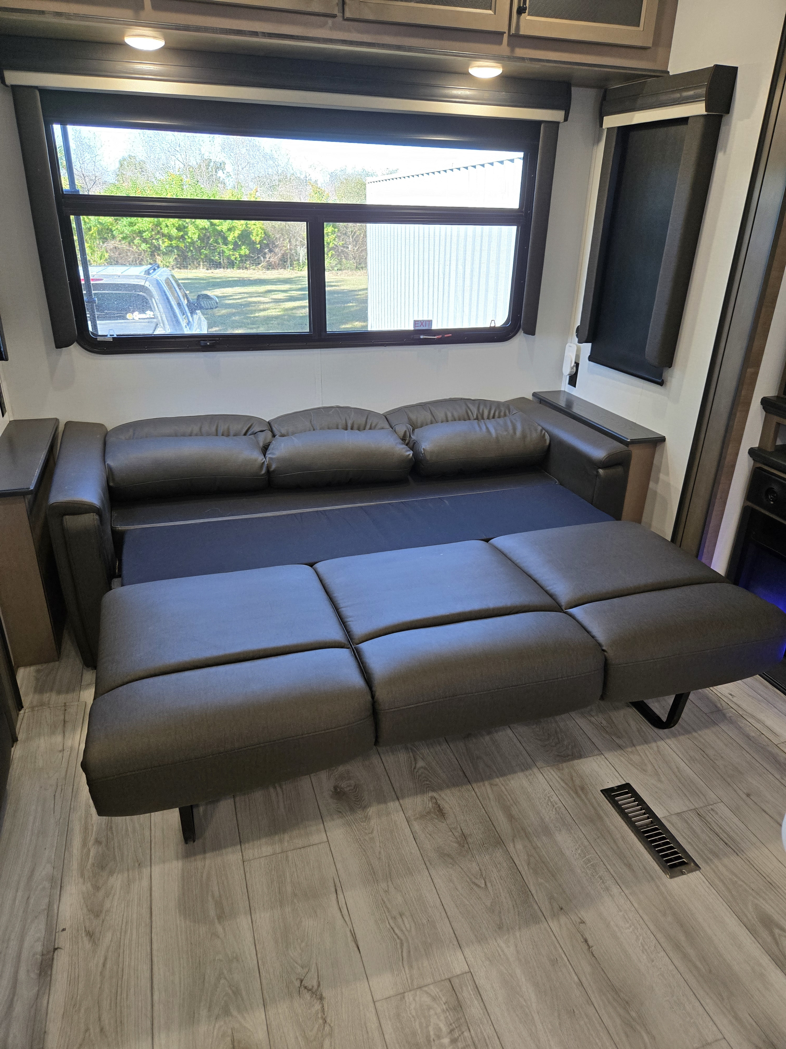 RV rental interior: a pull-out sofa bed in a modern living space with a large window overlooking trees and a building.. Lazy Days Keystone Montana 2024